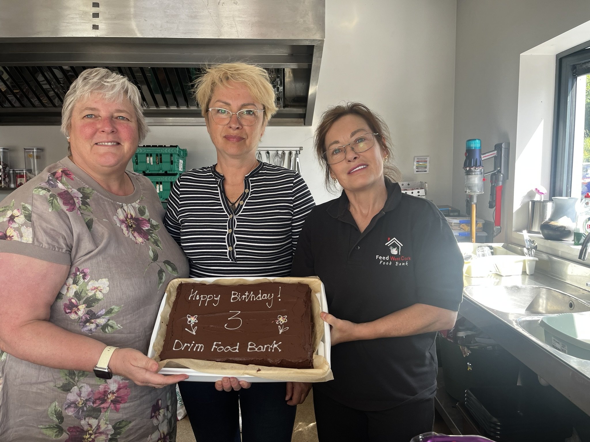 Three women standing in a kitchen, holding a birthday cake with chocolate frosting that has a message written on it. The cake reads 'Happy Birthday! 3 Drum Food Bank.' The women are smiling and looking at the camera.
