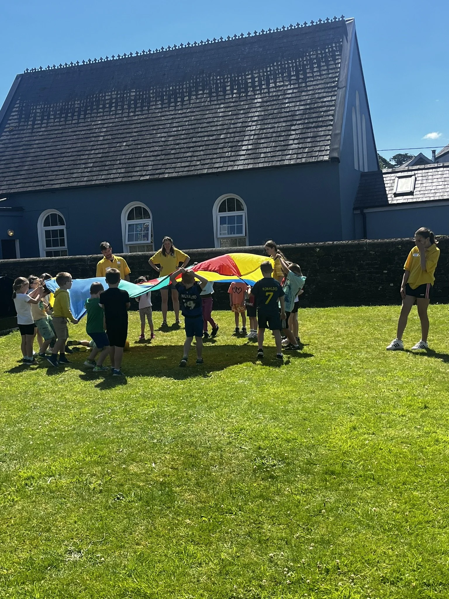Children playing with a colorful parachute outdoors on a sunny day, supervised by adults, in front of a blue building with arched windows and a sloped roof.