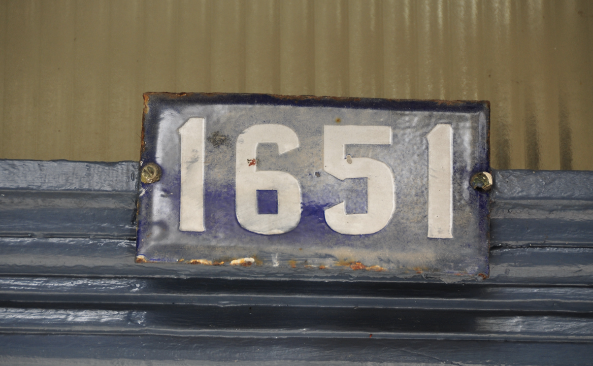 Weathered metal address plate with the number 1651 attached to a dark gray building exterior.