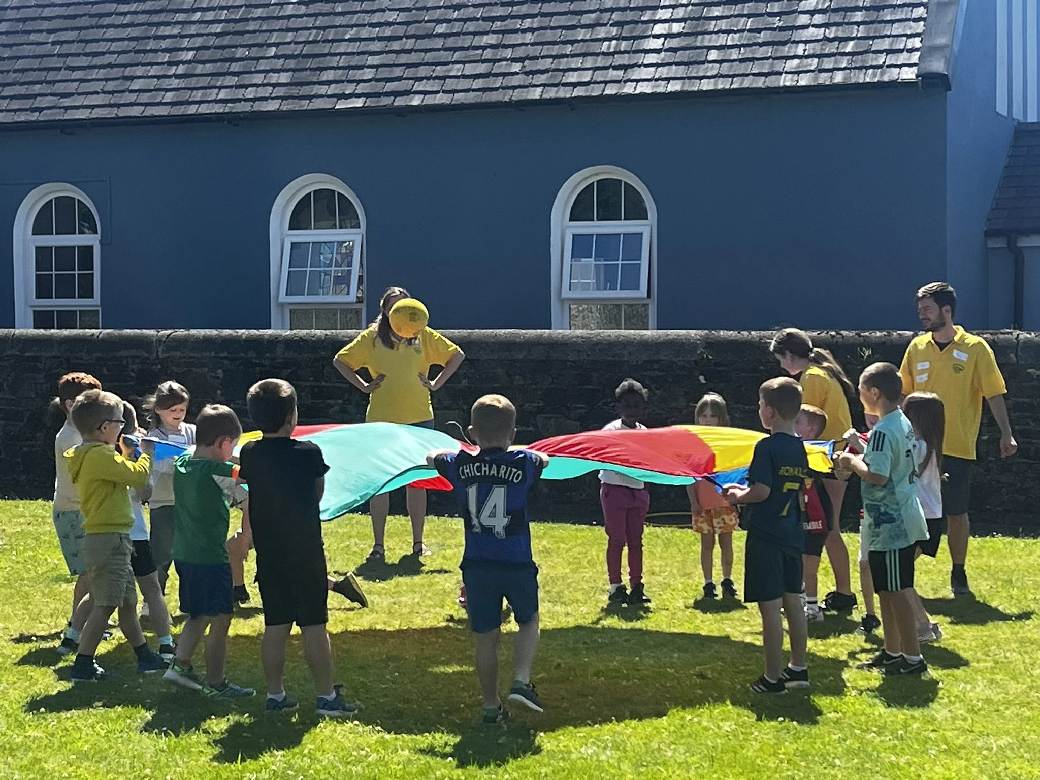 Children and adults by a stone wall holding a large multicolored parachute, in front of a blue building with arched windows.