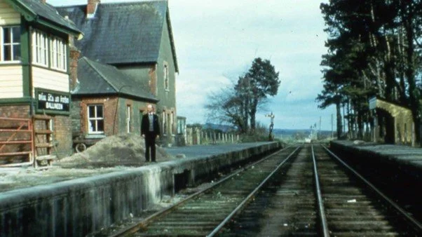 A man in a suit standing near train tracks next to a small station building with trees and a rural landscape in the background.