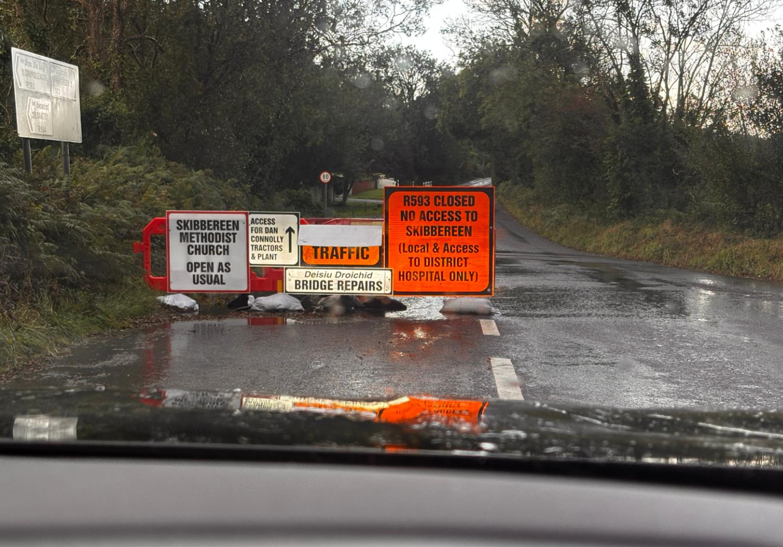 Roadblock with multiple signs indicating R593 is closed, with restricted access only for district hospital and local residents, wet road surface, rain droplets on windshield, and trees lining the road in the background.