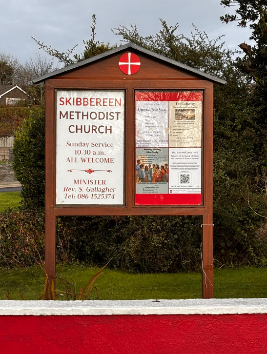 A wooden church notice board for Skibbereen Methodist Church displaying service times and contact information, with a small circular symbol featuring a red cross at the top, set against trees and a cloudy sky.