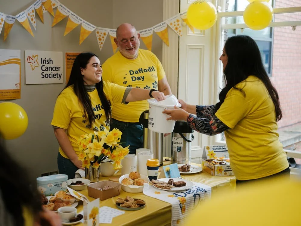 People at a fundraising event for Daffodil Day, organized by the Irish Cancer Society. Three individuals wearing yellow shirts are smiling; one is serving hot beverages to another person. The table is decorated with yellow balloons, flowers, and various baked goods. There are signs and banners supporting Daffodil Day.