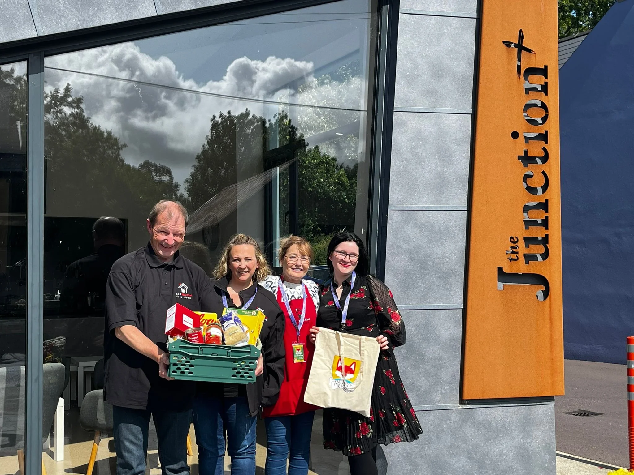 Four people standing outside the entrance of 'The Junction' restaurant, holding gift baskets and a tote bag, smiling for the camera on a sunny day.
