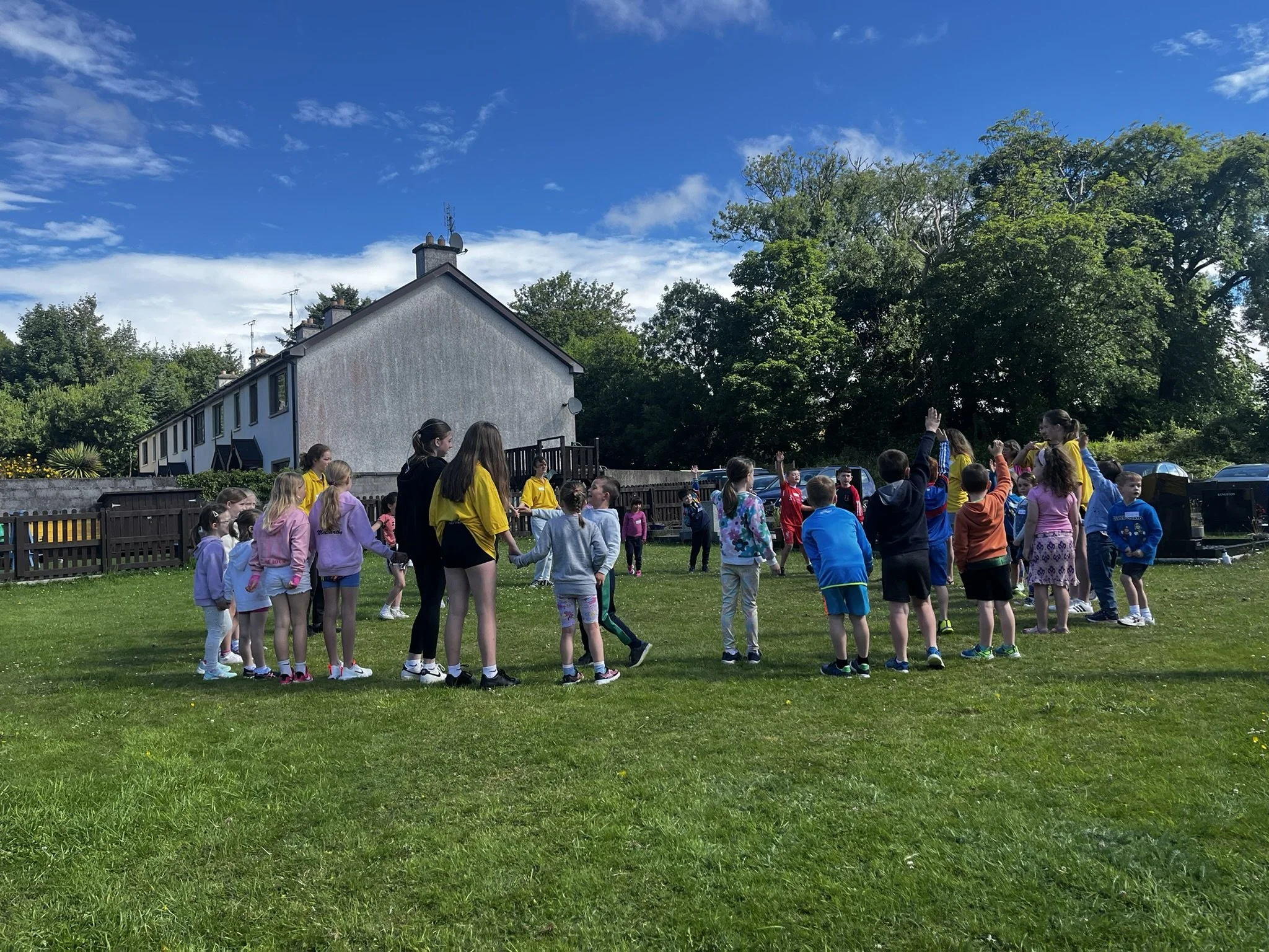 Children playing outdoors on a grassy field under a blue sky with scattered clouds, some of them holding hands and others raising their hands, with a house, trees, and parked cars in the background.