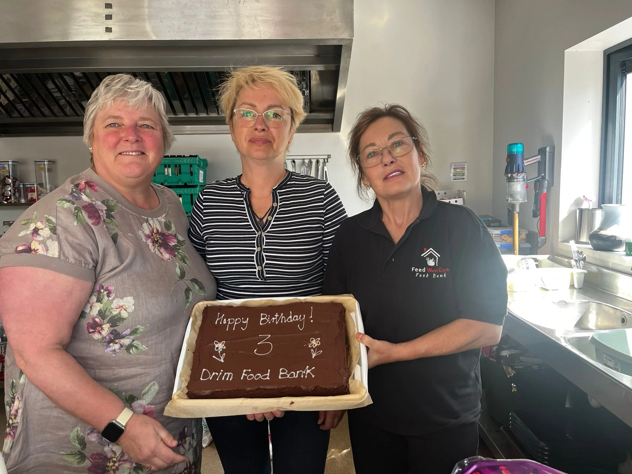 Three women in kitchen holding a birthday cake with message for Drum Food Bank.