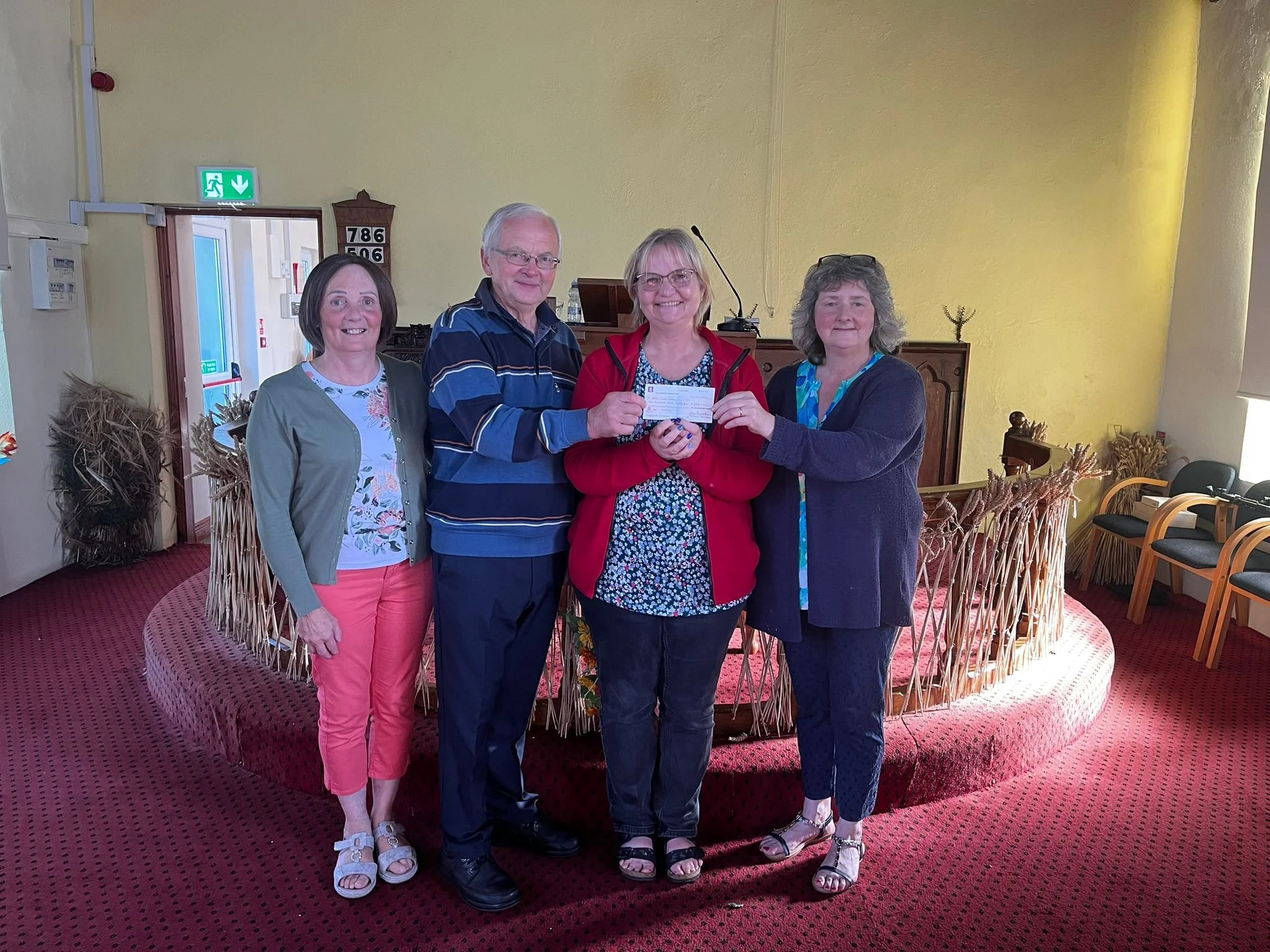 Four people standing indoors on red carpeted floor, holding a check, and smiling at the camera. Behind them is a wooden altar with floral decorations, and to the right, a large window partially covered by a blind.
