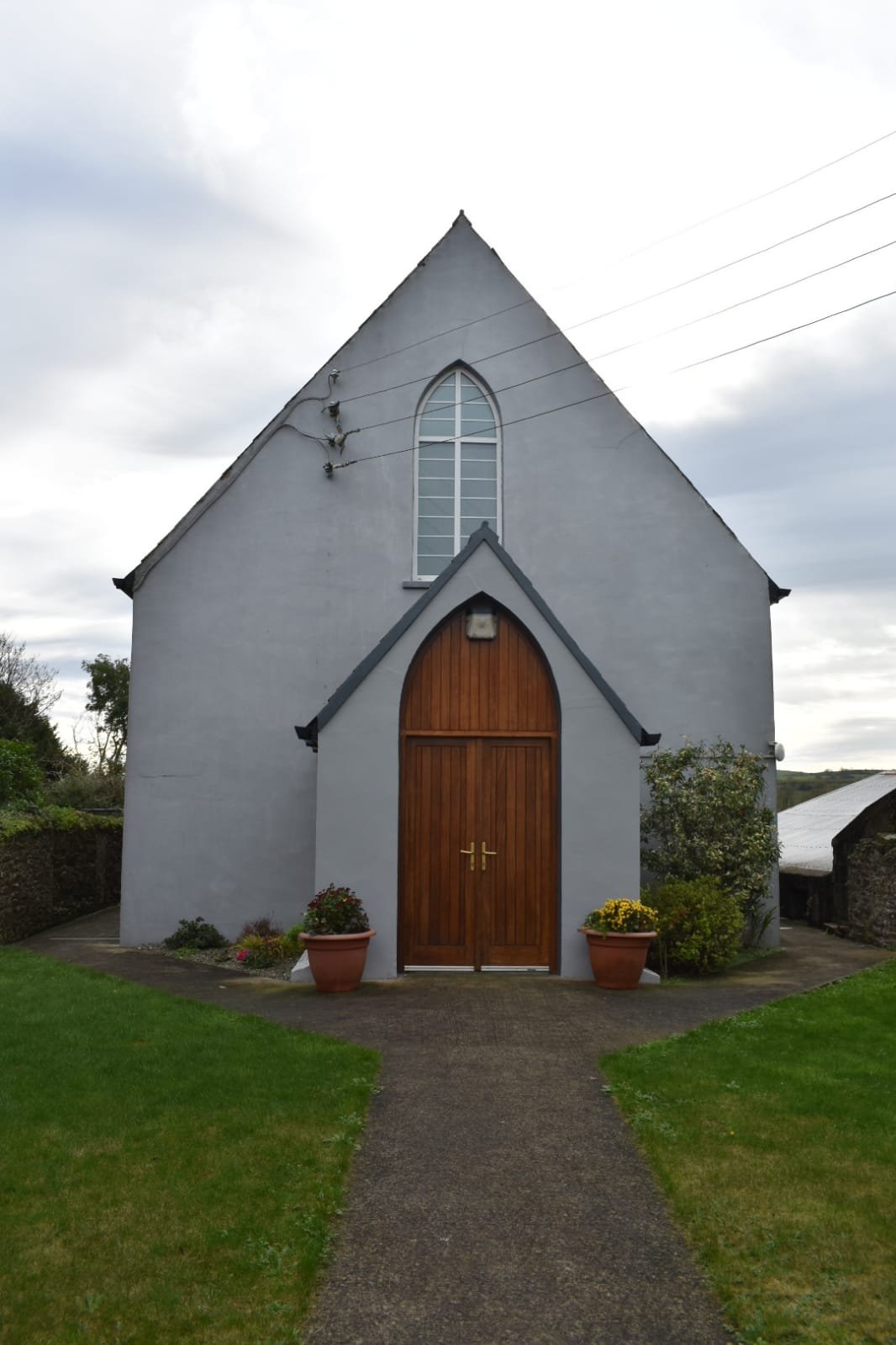A small gray church with pointed roof and wooden doors, surrounded by green lawn, potted plants, and cloudy sky.