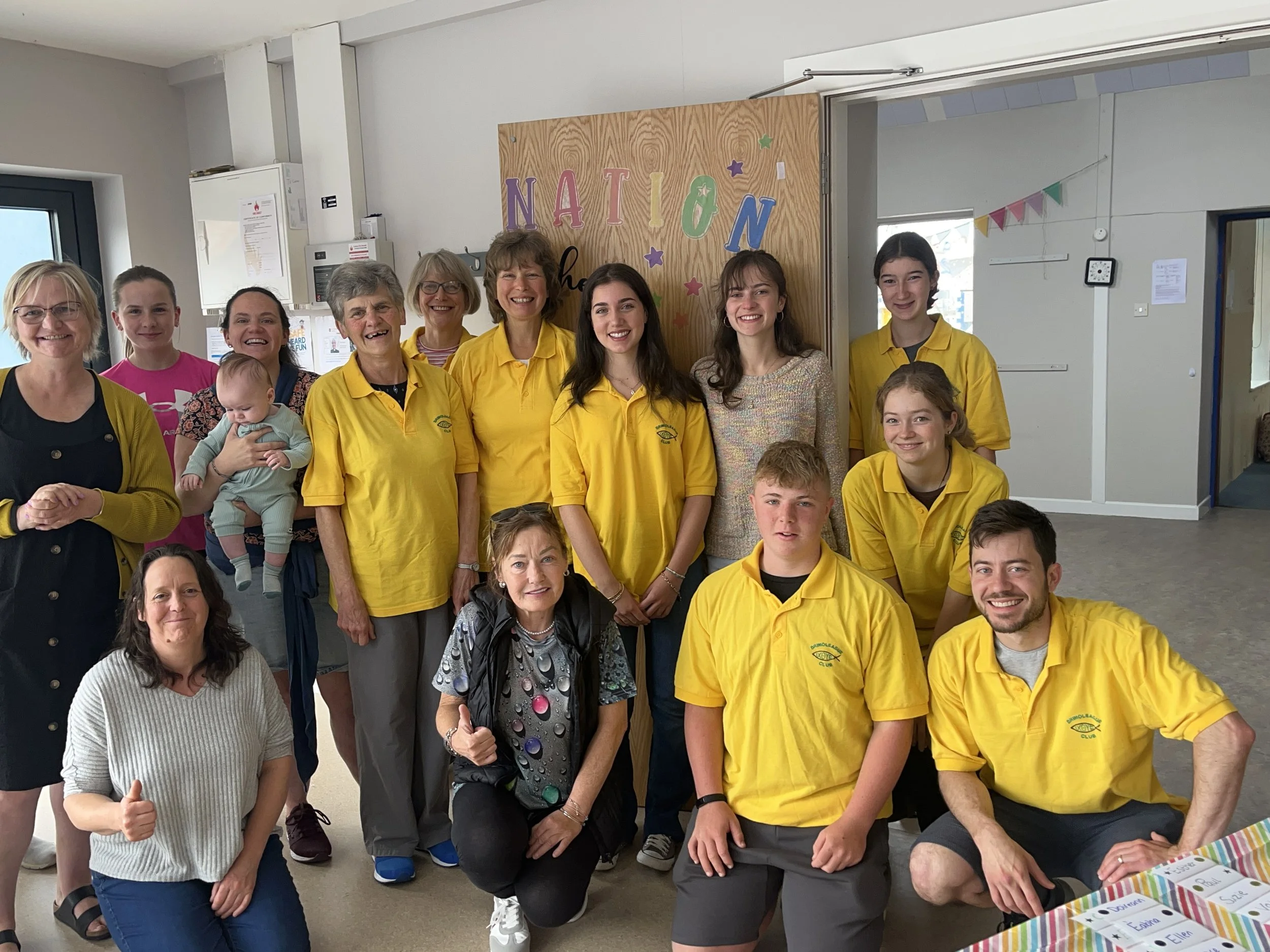 Group photo of children, teenagers, and adults standing indoors, some wearing yellow shirts with a logo, in front of a decorated wooden board that says 'NATION'.