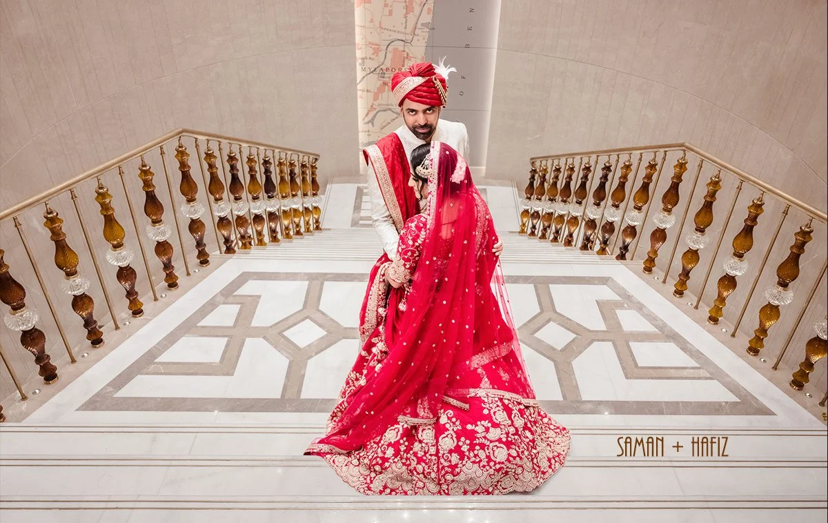 A Muslim bride and groom in traditional Indian wedding attire on a marble staircase with gold and brown railing. The bride wears a red embroidered lehenga with a sheer dupatta, and the groom wears a white sherwani with a red and gold turban.