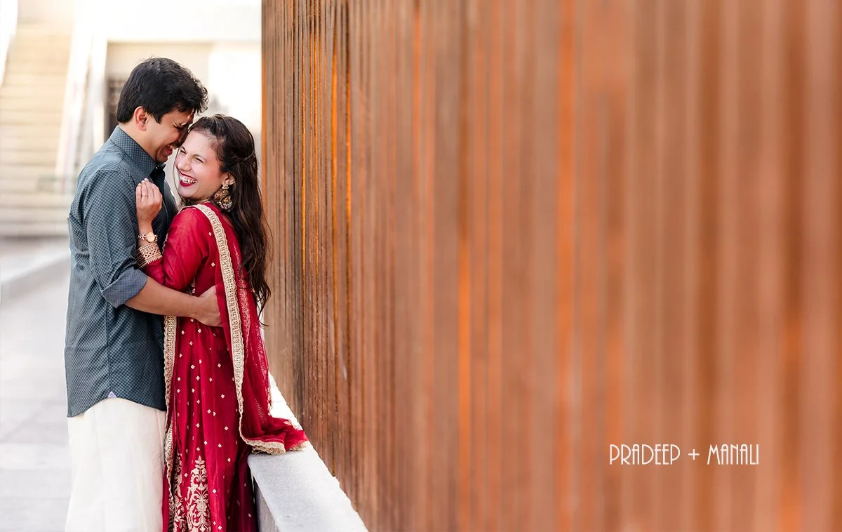 A couple sharing a joyful moment outdoors, standing close together near a wooden fence. The woman is wearing a red traditional outfit with gold embroidery and jewelry, while the man is wearing a dark shirt and light-colored pants. They are smiling and laughing, their foreheads and noses touching in a happy embrace.