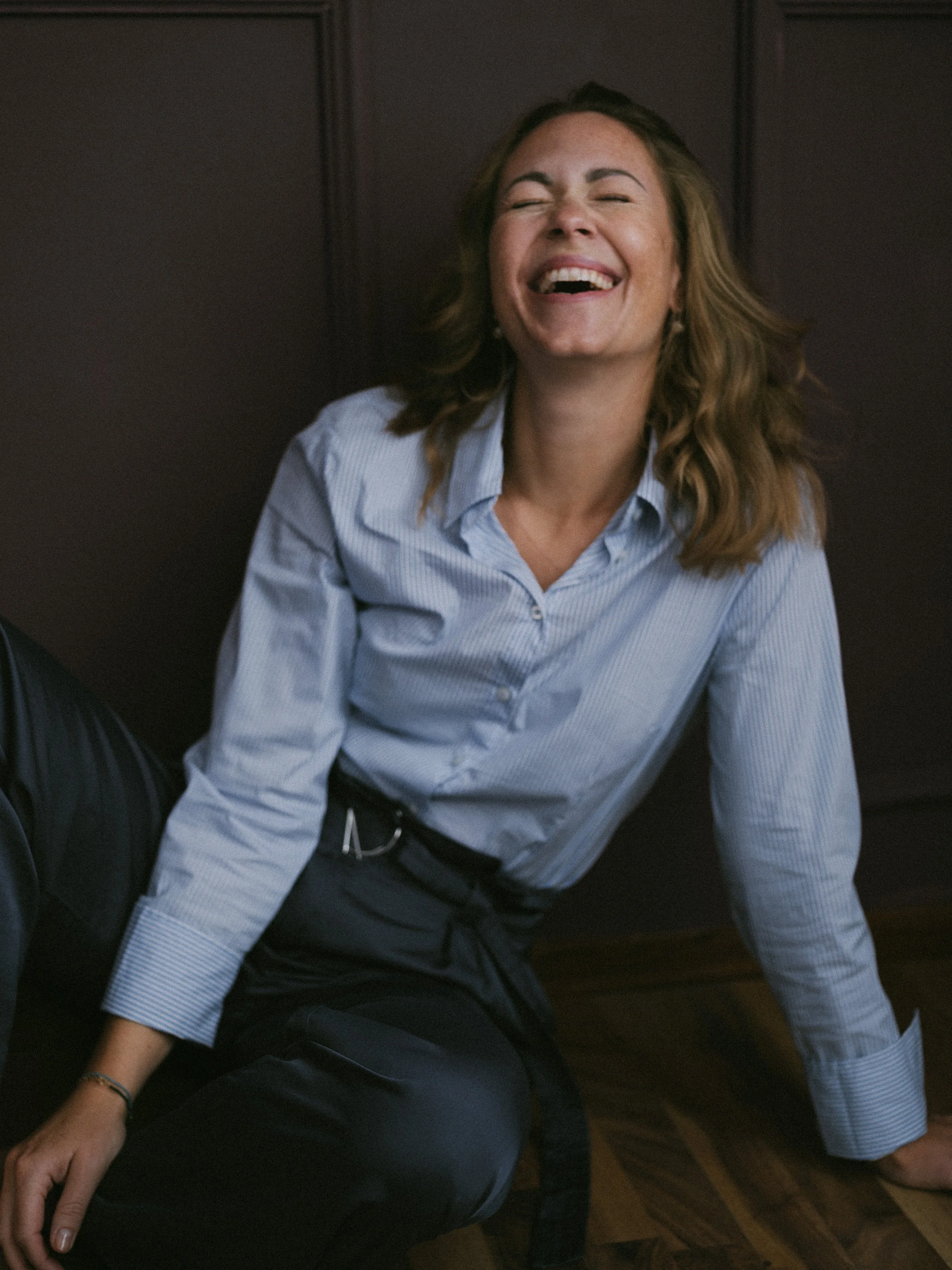 Frau in blauer Hemd sitzt lachend auf dem Boden, vor einer dunklen Wand.