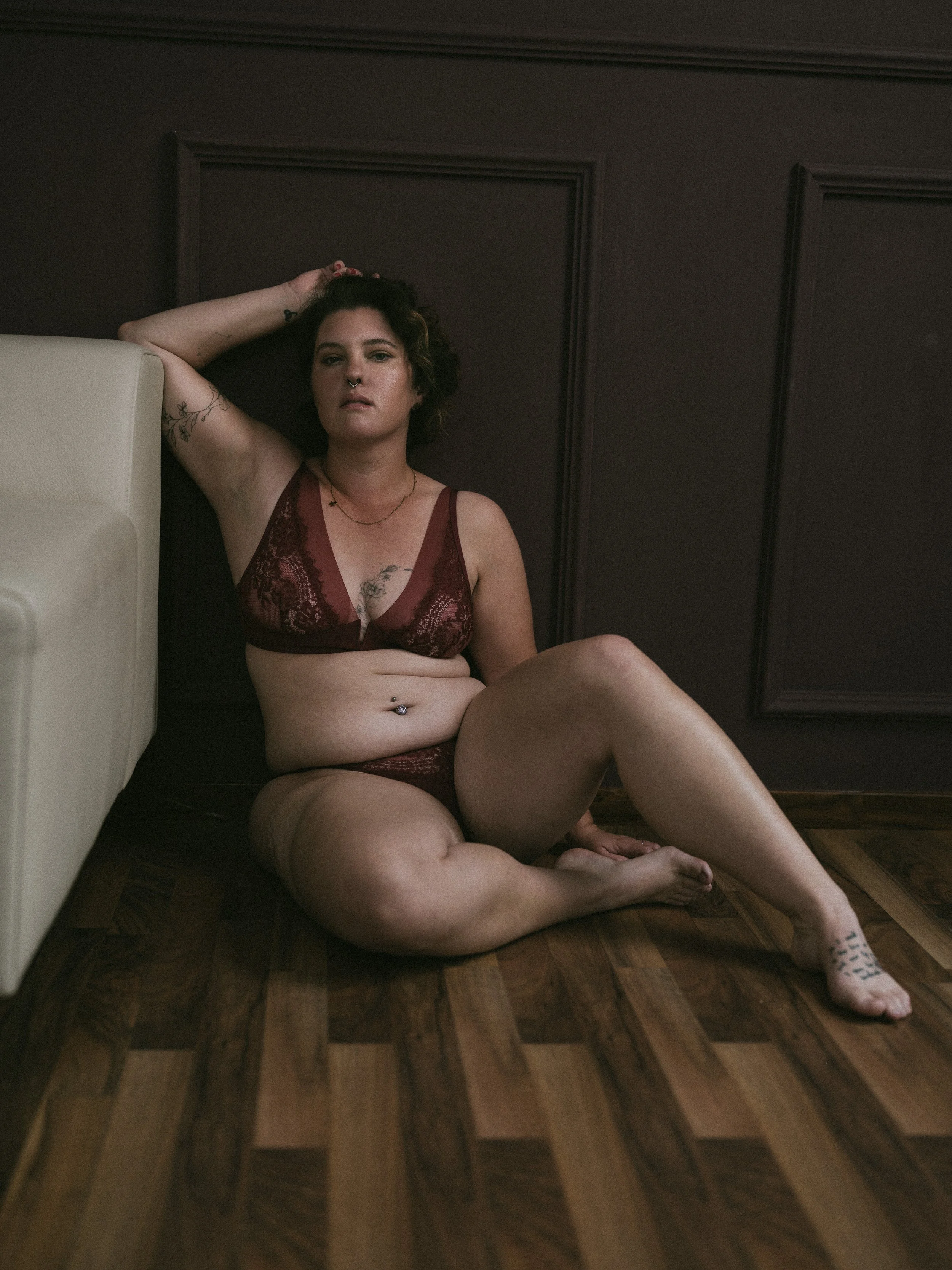 Eine Frau sitzt auf einem Holzboden vor einer dunkel gestrichenen Wand, trägt rote Spitzenwäsche, hat Tattoos und Piercings, und blickt direkt in die Kamera.