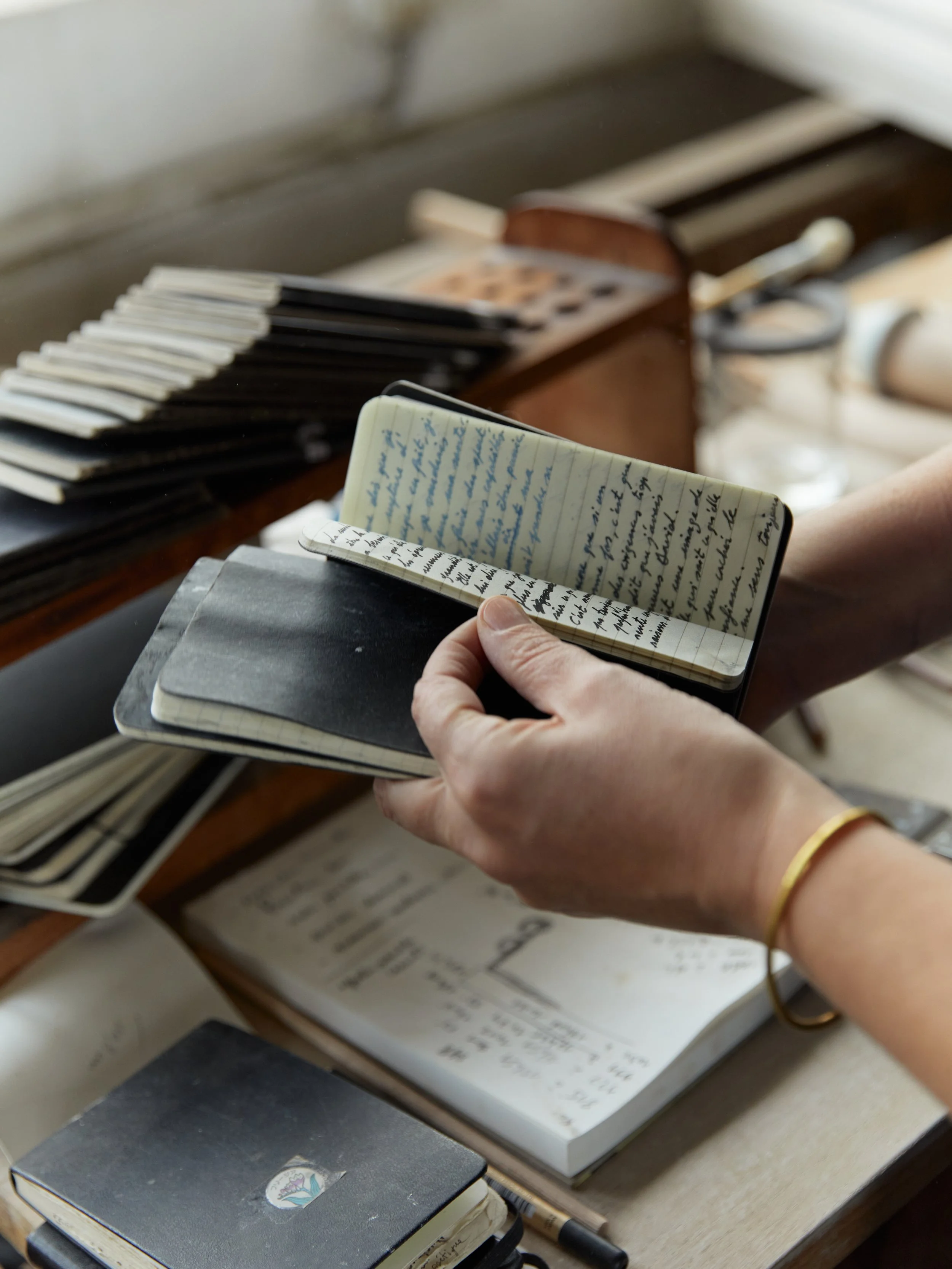 Une personne tient un petit carnet ouvert avec des écritures manuscrites, à côté de plusieurs livres empilés sur une table, avec un fond flou de plus de livres.