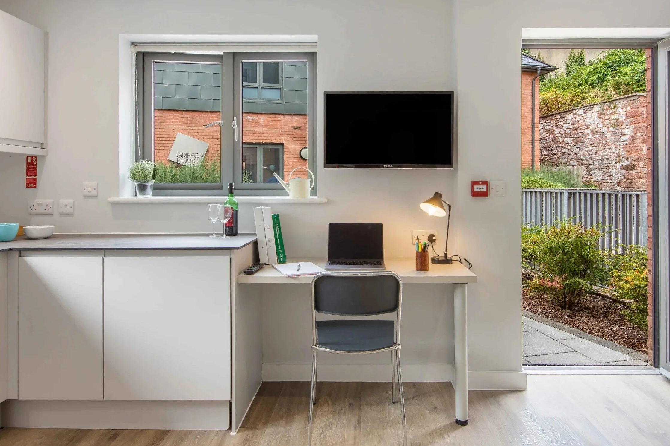 A bright, modern kitchen and workspace with white cabinets, a window showing a brick building outside, a small desk with a laptop, a lamp, and office supplies, and an open door leading to a small outdoor patio with greenery.