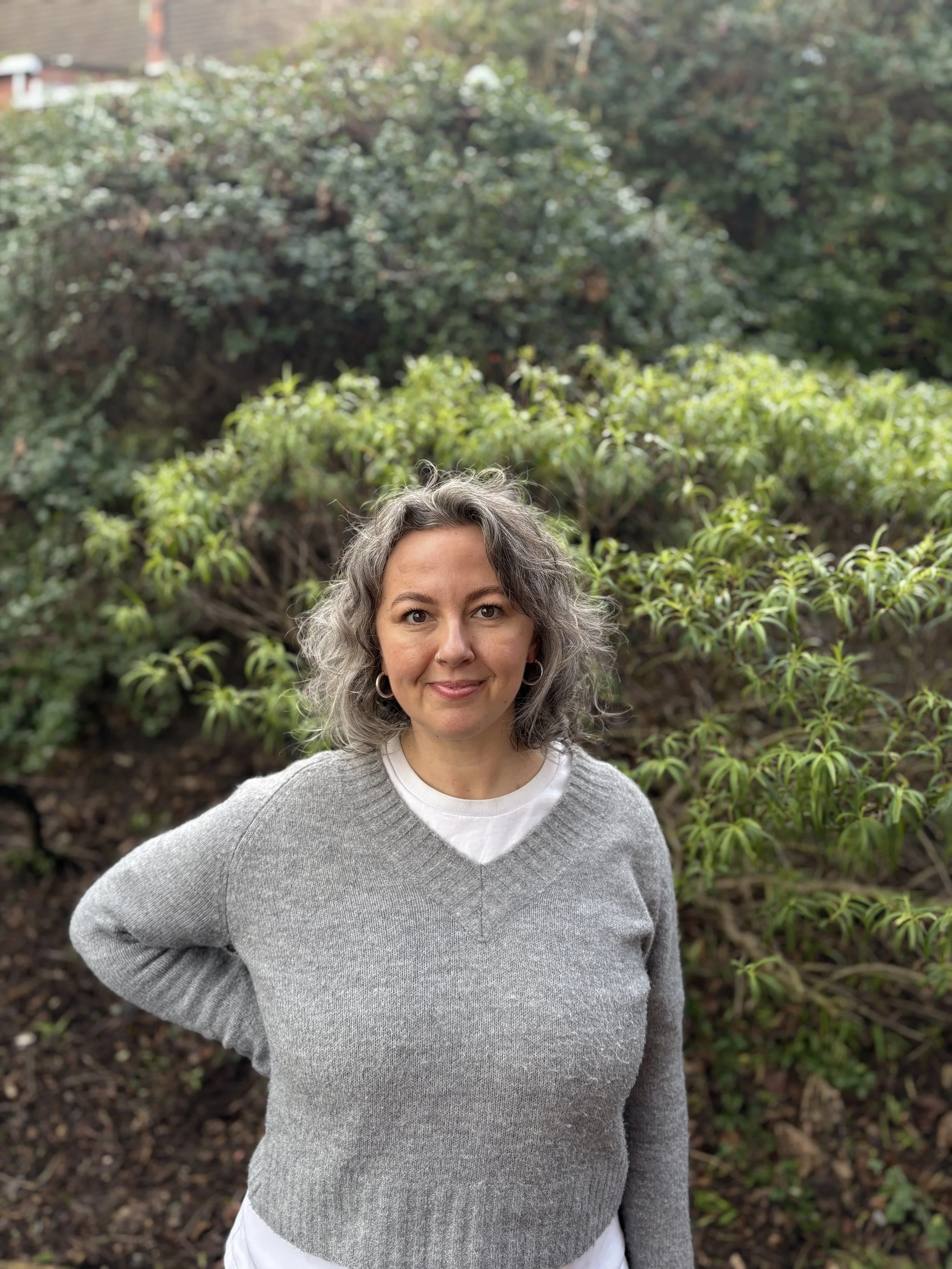 A woman with long brown hair smiling outdoors in front of lush green plants and white flowering bushes.