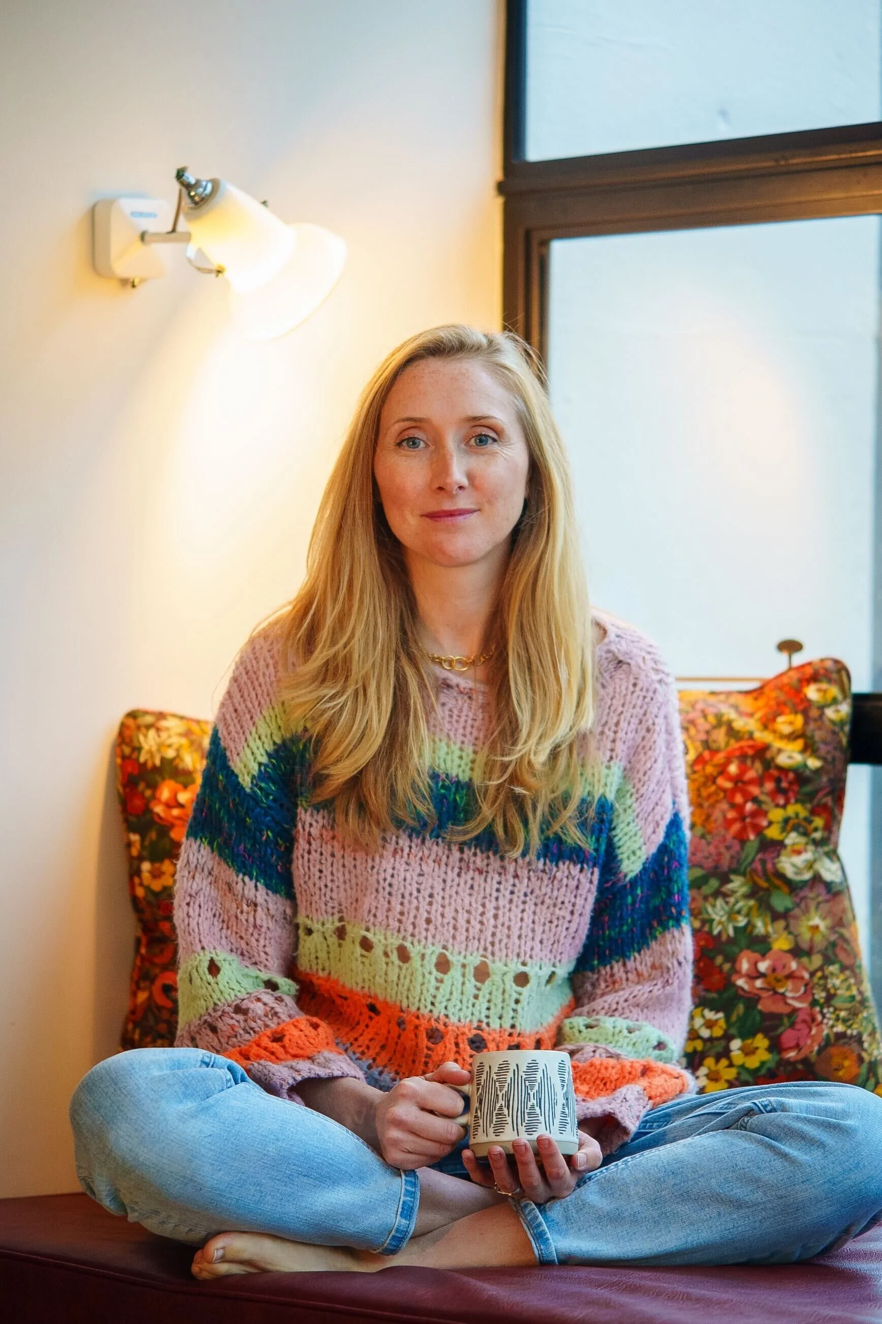 A woman with long blonde hair, wearing a colorful knitted sweater and jeans, seated cross-legged on a cushioned window seat, holding a mug with a black and white pattern, near a window and a wall-mounted light.