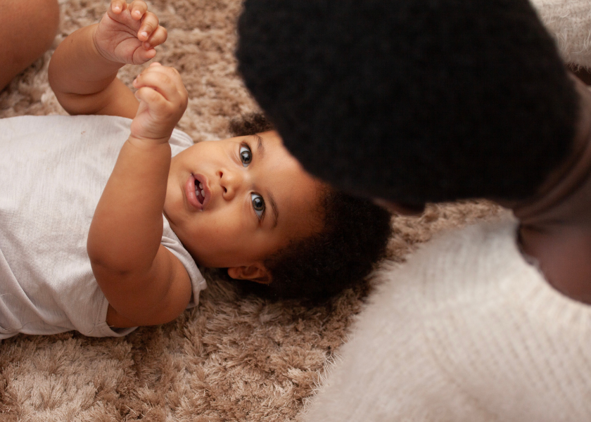 A baby lying on a brown carpet, looking up at an adult with short hair and a beard, engaging in eye contact.
