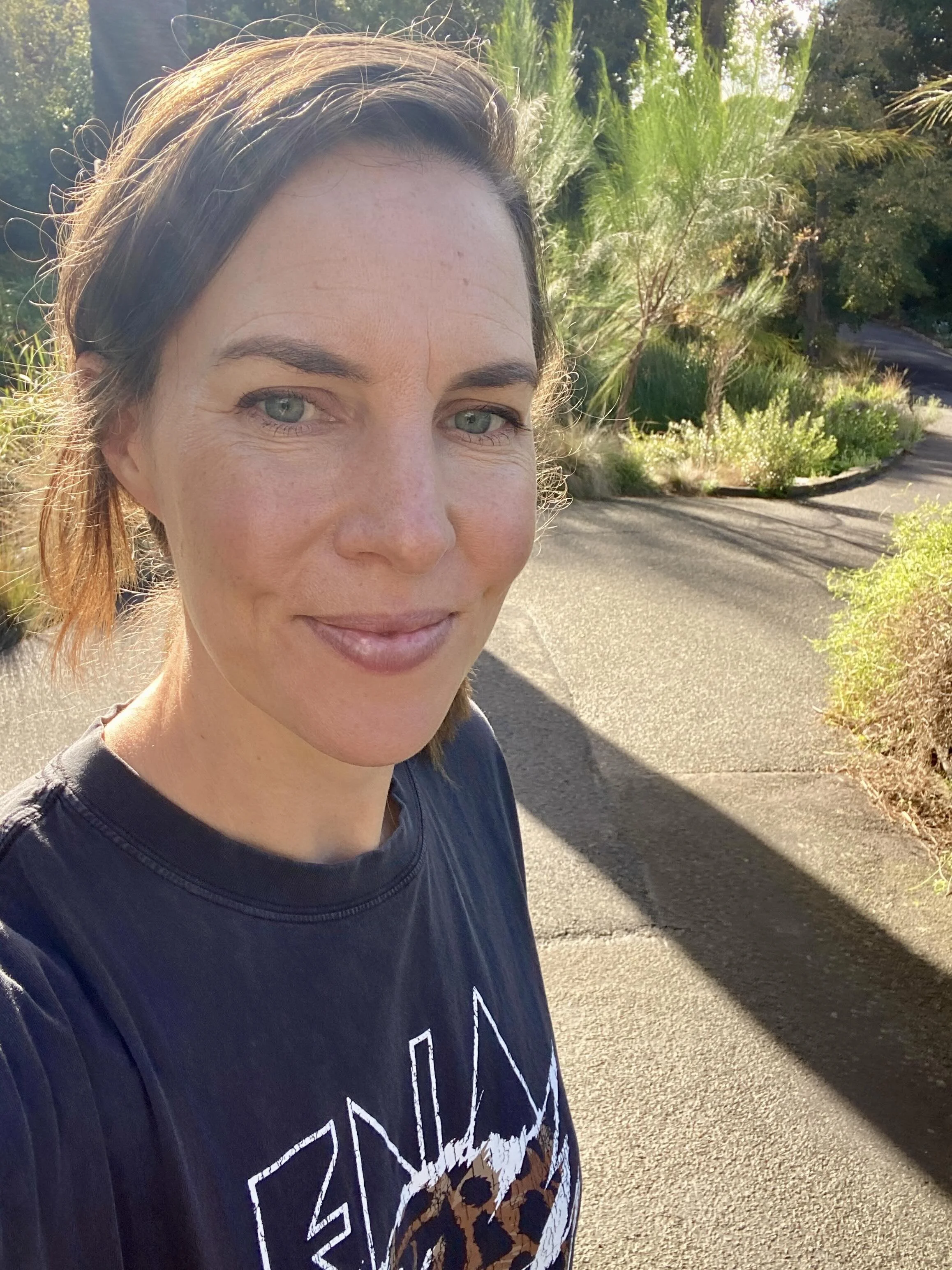 Close-up of a smiling woman with blue eyes and brown hair, standing outdoors on a sunny day, with trees and plants in the background.
