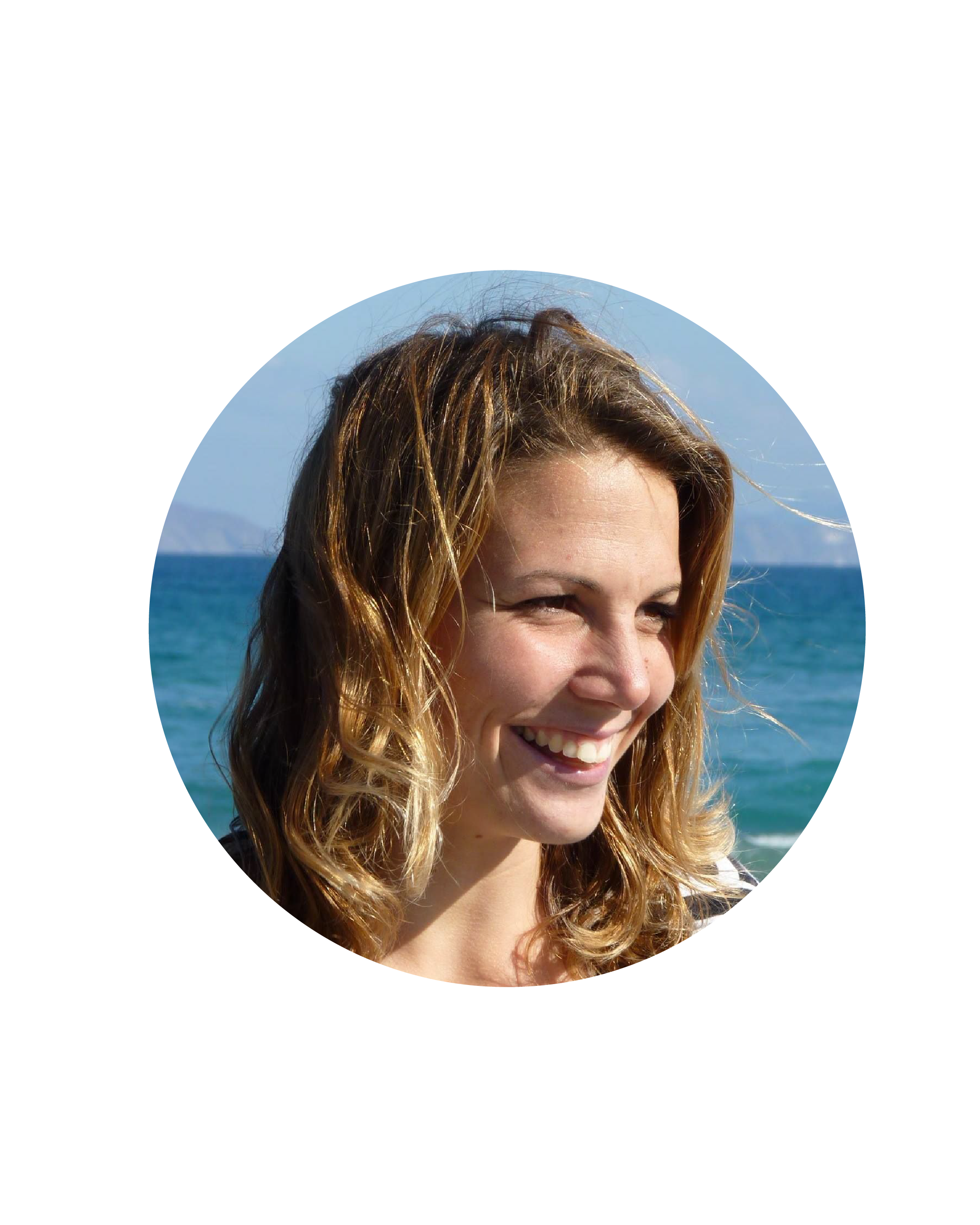 A smiling woman with wavy, shoulder-length brown hair standing at the beach with ocean and mountains in the background.