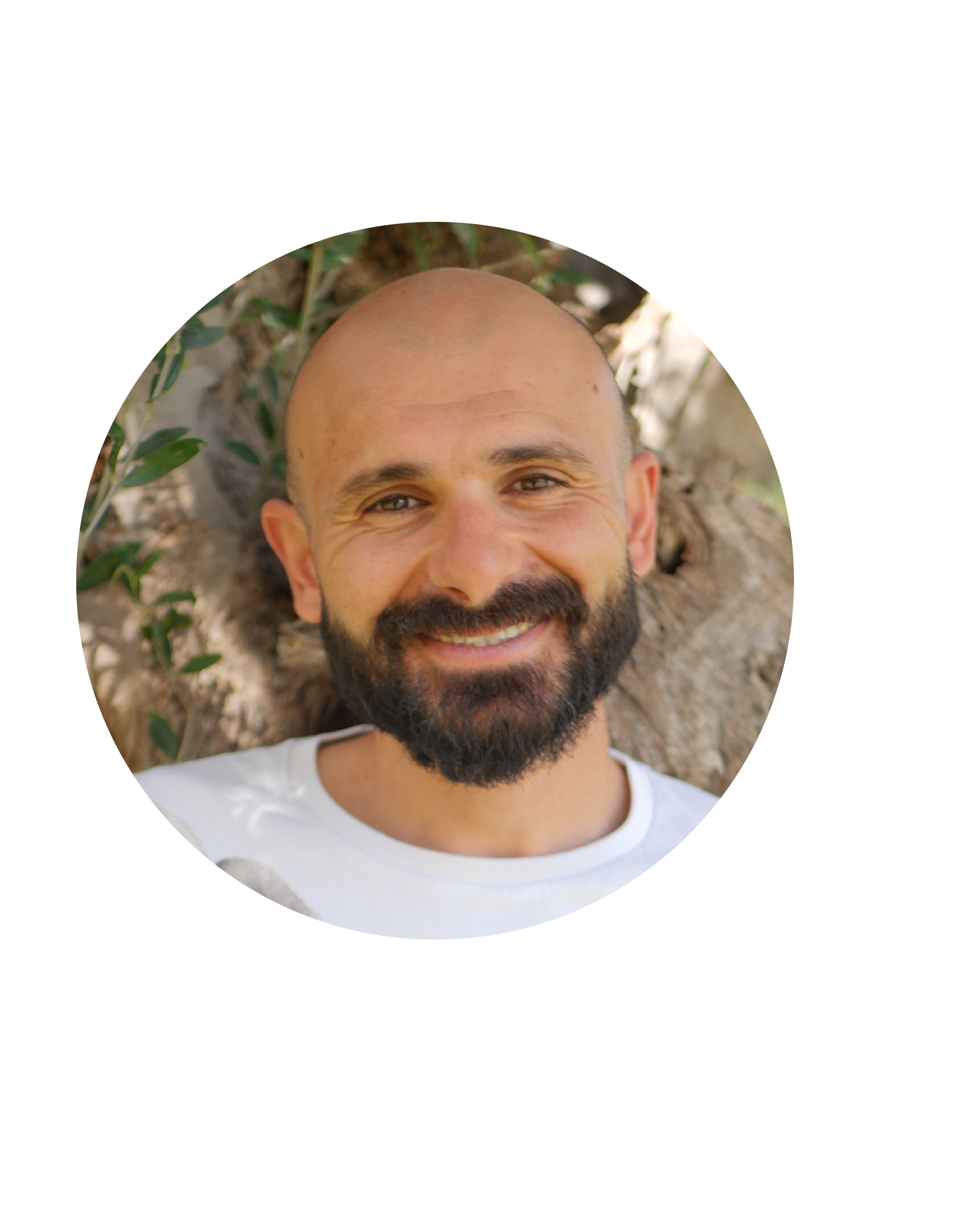 A smiling man with a beard and a shaved head, wearing a white shirt, leaning against a tree outdoors with green leaves.