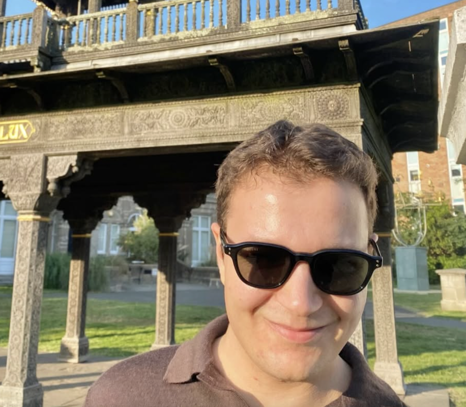 Young man with short brown hair wearing black sunglasses and a dark brown shirt, smiling outdoors in sunlight, standing in front of an ornate, vintage metal gazebo with decorative carvings, set in a park with grass and trees in the background.