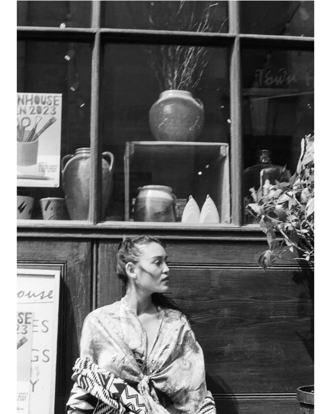 A woman seated against a wooden wall, wearing a patterned shawl, with a wooden shelf filled with vases, plates, and decorative items behind her.