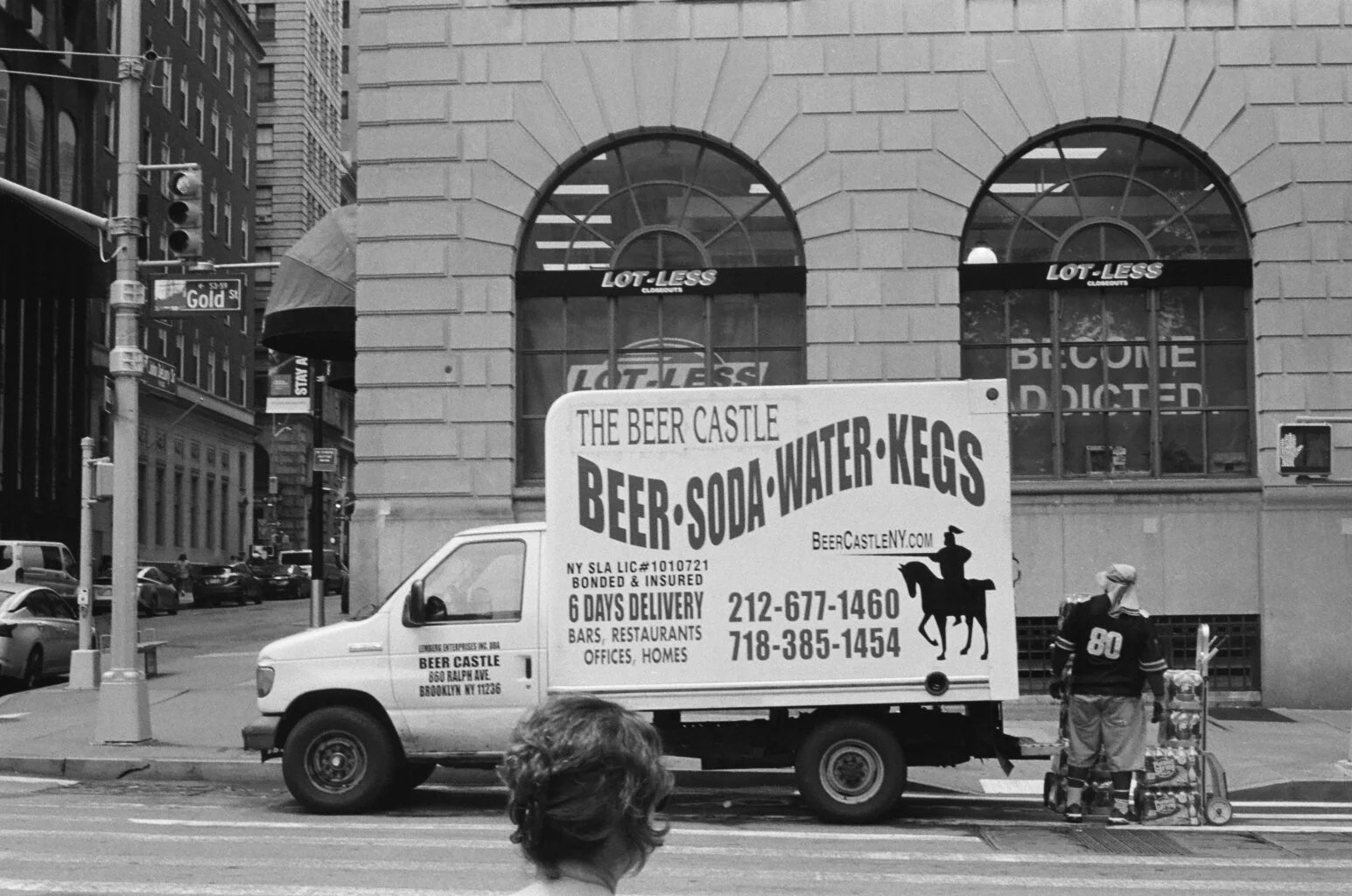 A street scene with a moving truck advertising The Beer Castle, which sells beer, soda, water, and eggs, with contact information and delivery details. There are pedestrians on the sidewalk, and tall buildings in the background. The truck has a billb
