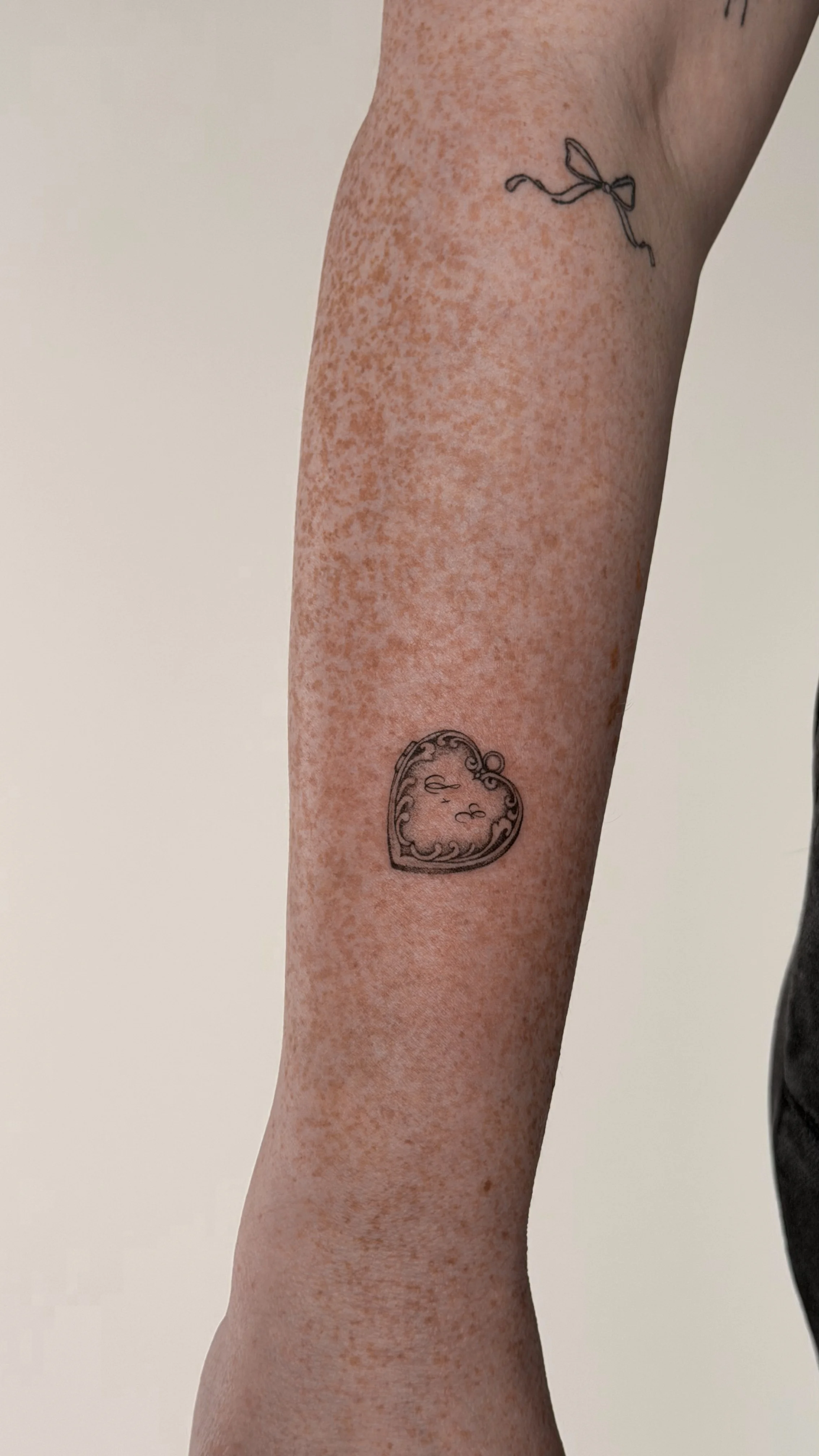 Close-up of a person's arm with two tattoos: a small bow near the shoulder and a heart-shaped locket with ornate detailing lower on the arm.