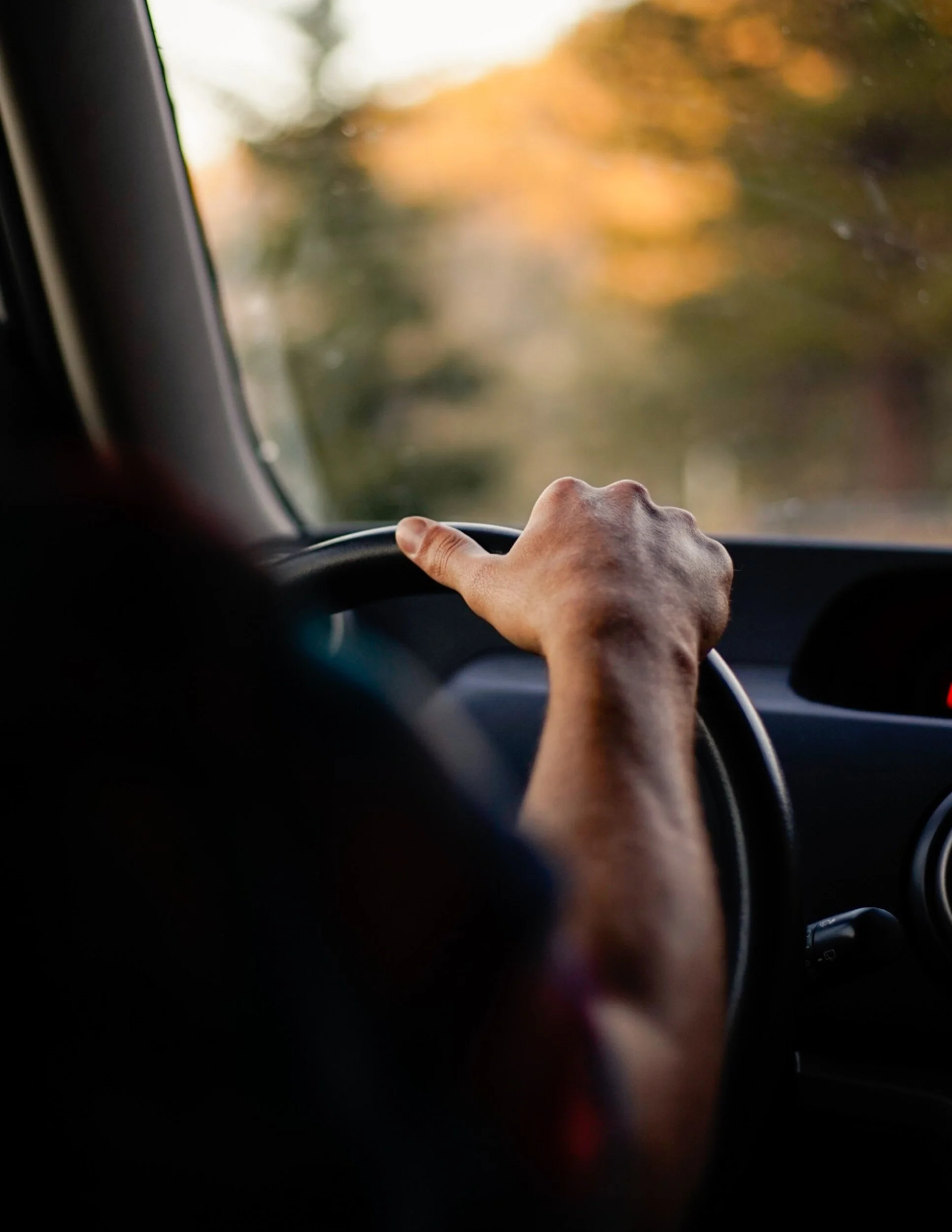 Person's hand on steering wheel inside a car with blurred outdoor background.