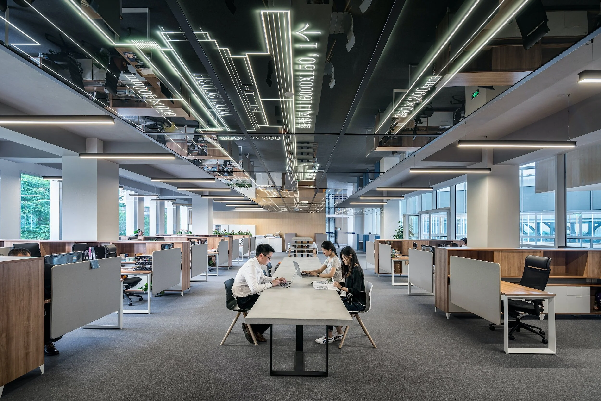 Open-plan modern office space with people working at a long table, cubicles on either side, and reflective ceiling panels showing digital designs.