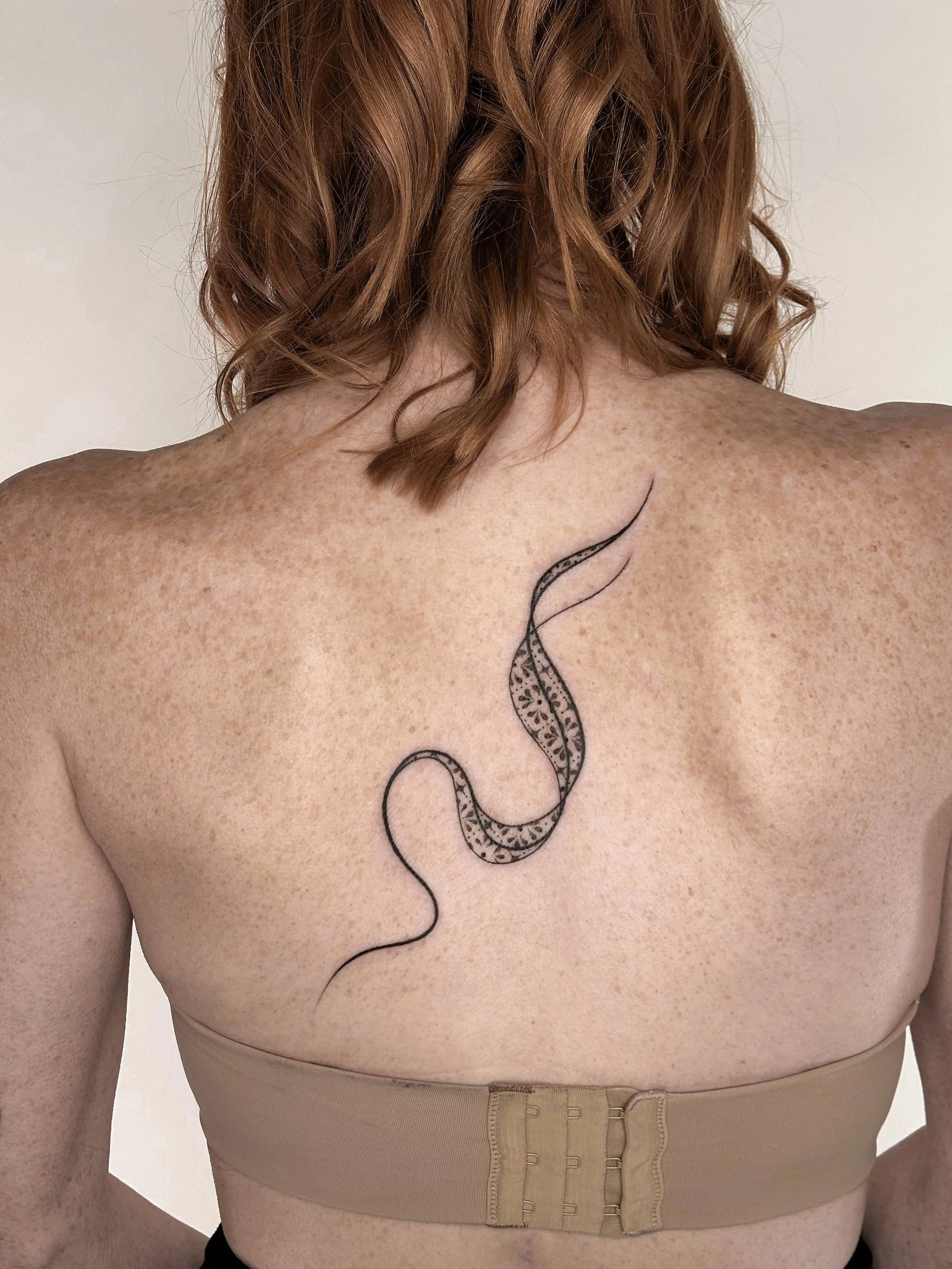 A woman with red hair and freckles showing a black ink snake tattoo on her upper back, just below her neck.