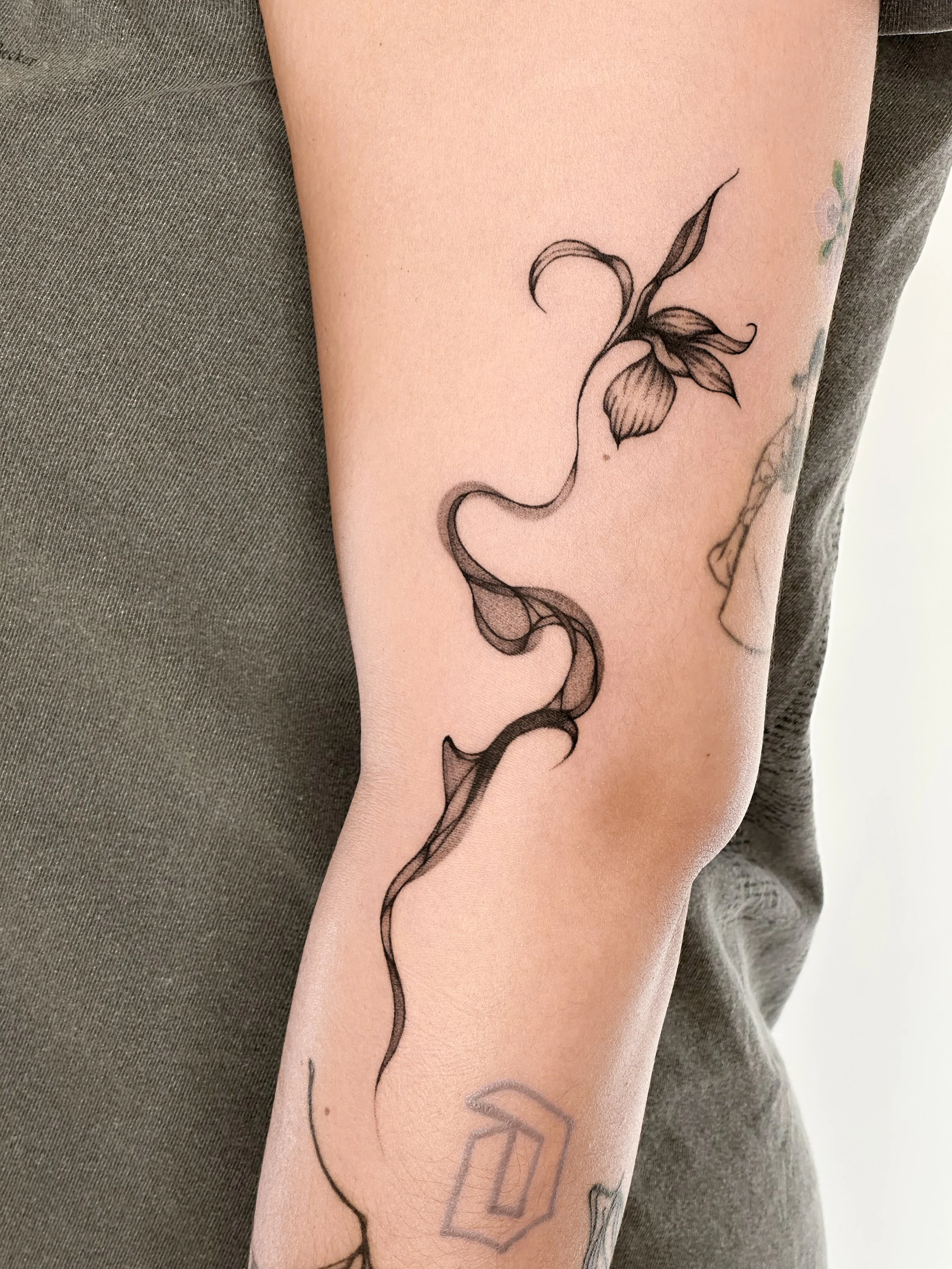 A black ink tattoo on a person's arm featuring a stylized, flowing flower with leaves and a long, swirling stem.