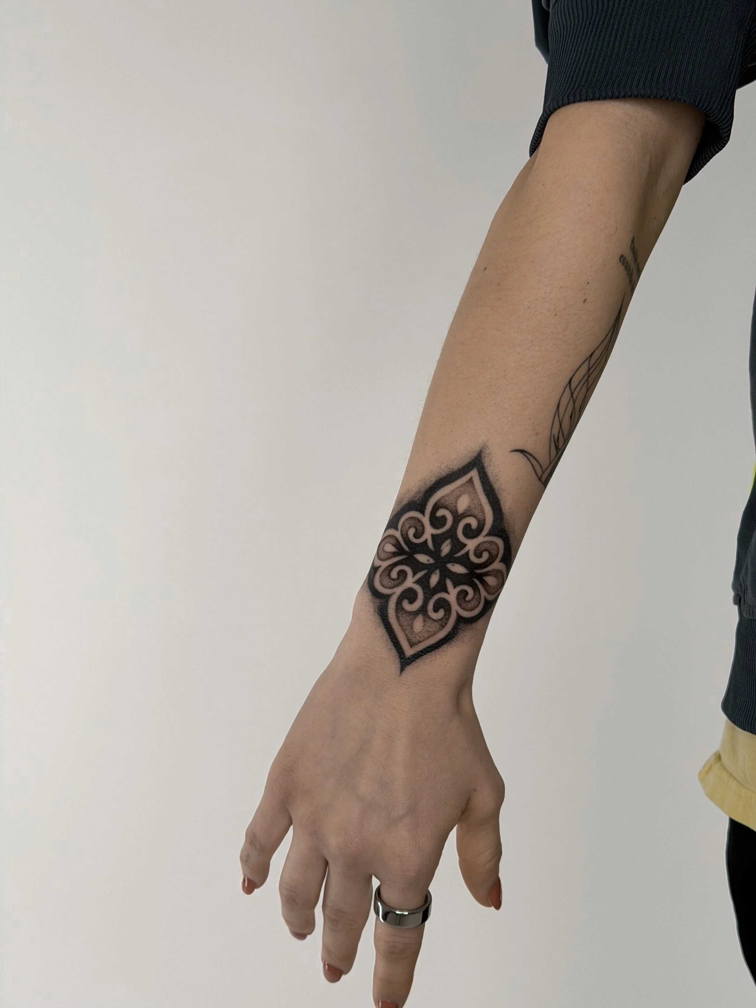 A person's arm extended downward, showing a black and pink tattoo with intricate, symmetrical design on the wrist.