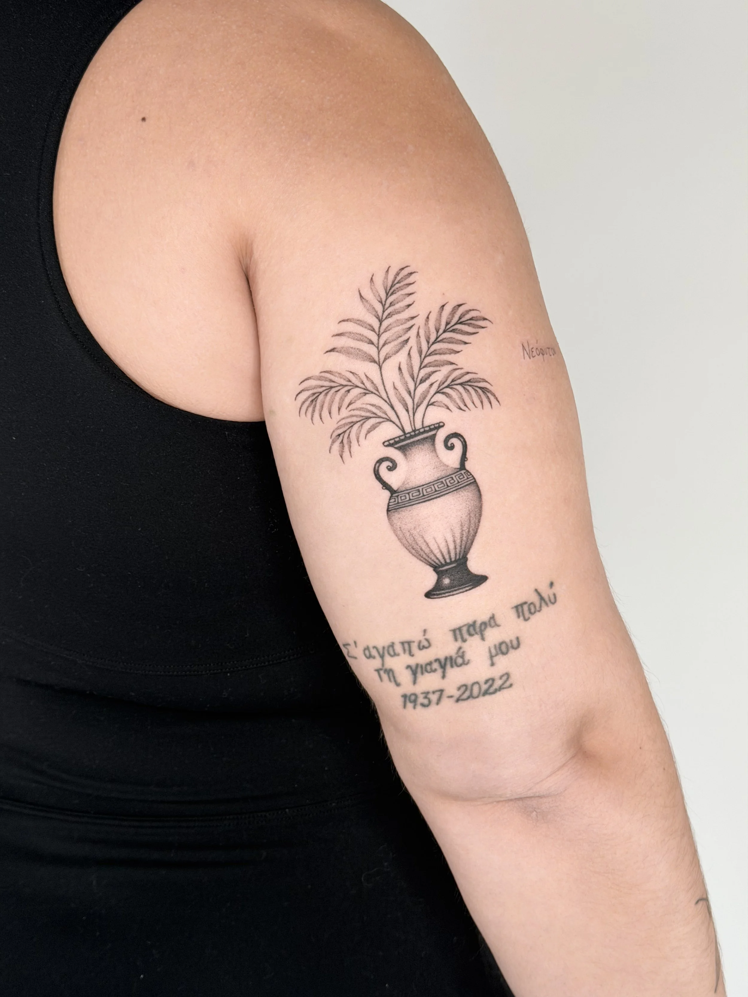 Tattoo of a decorative vase with leaves on the upper arm, with Greek text and dates below that read: 'I love you very much, my daughter, 1937-2022.'