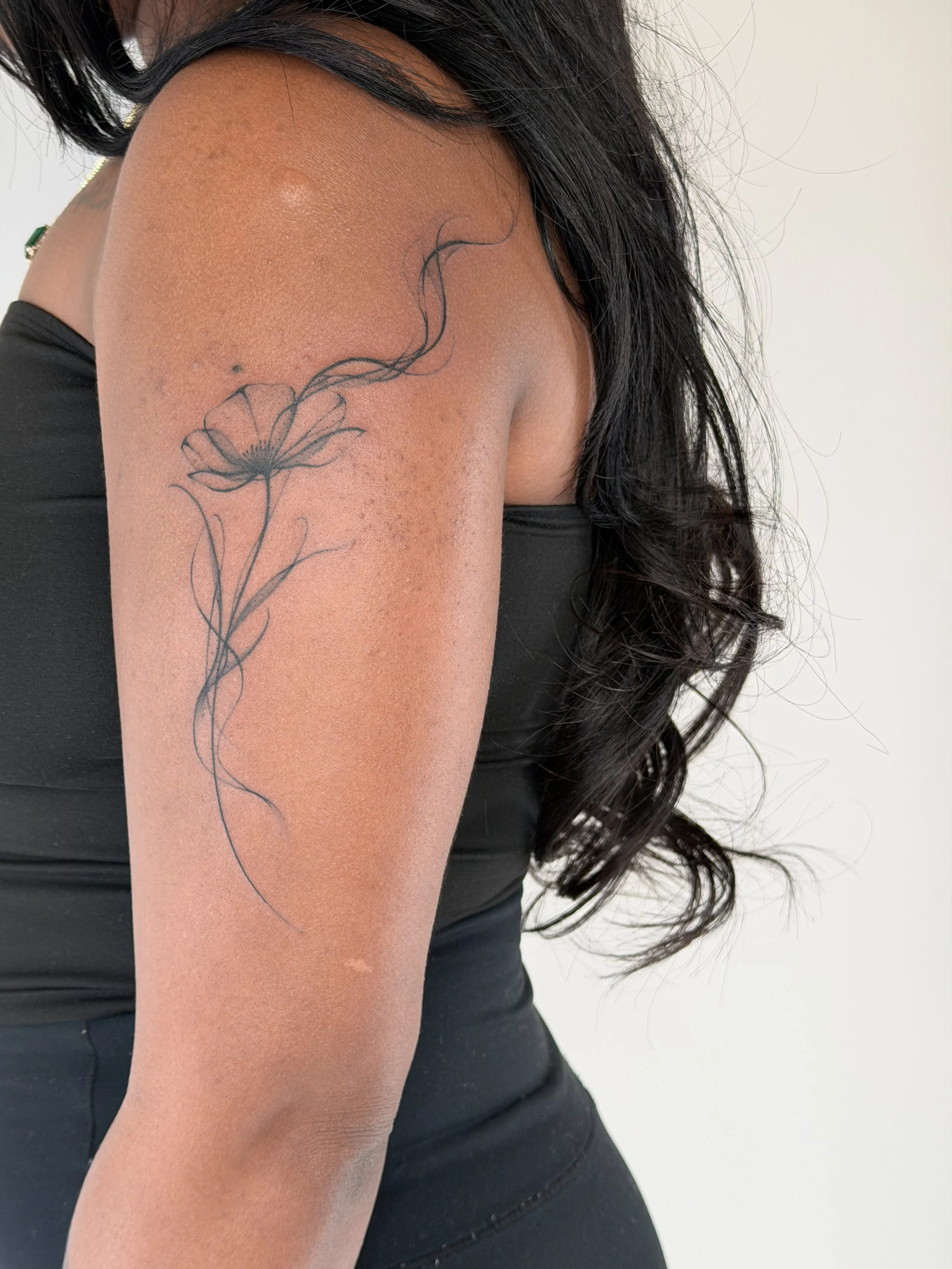 Close-up of a woman's upper arm with a delicate black flower tattoo, wearing a sleeveless black top, with long wavy black hair.