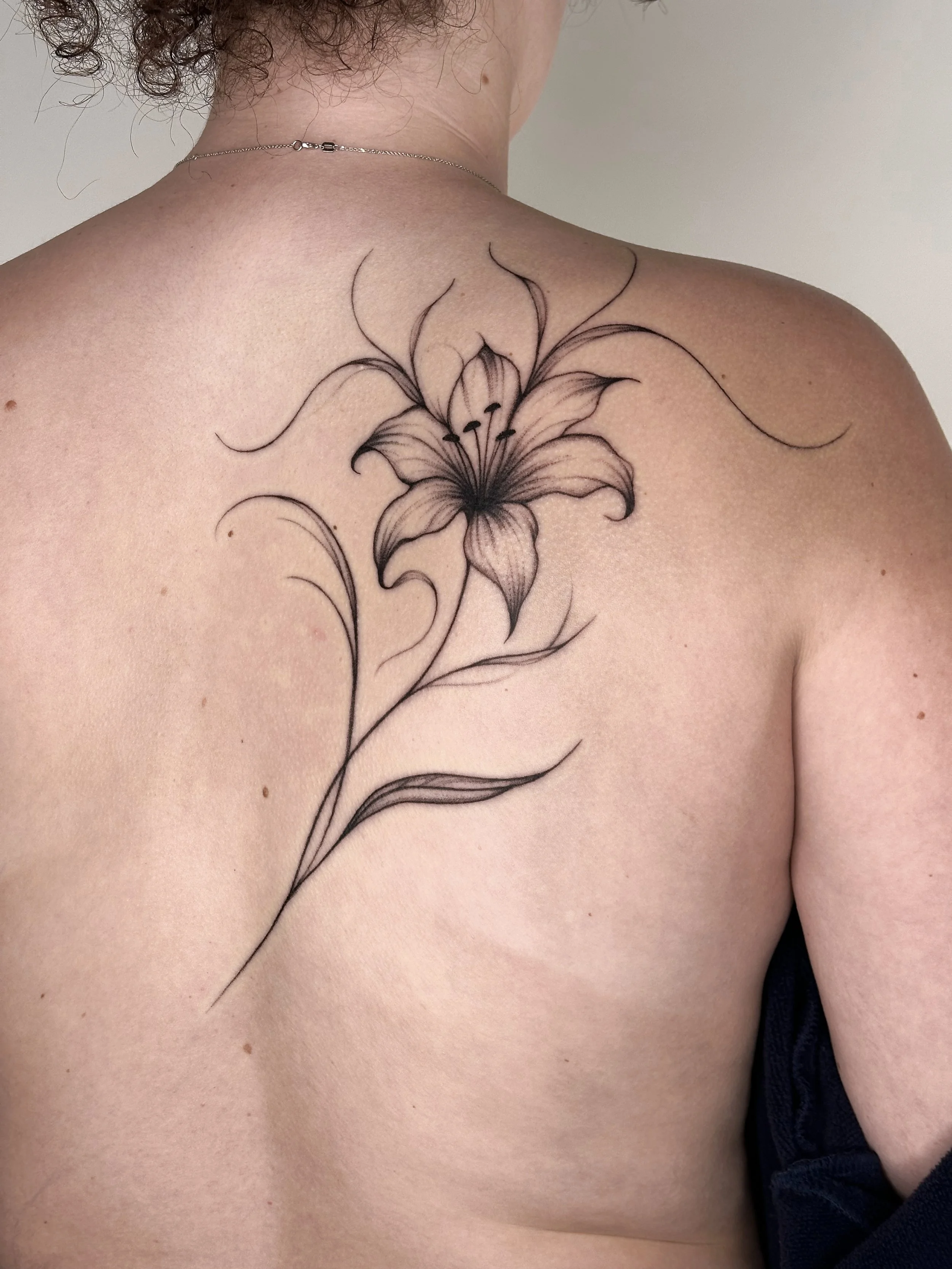 Black ink floral tattoo of a lily on upper left back and shoulder of a person with light skin.