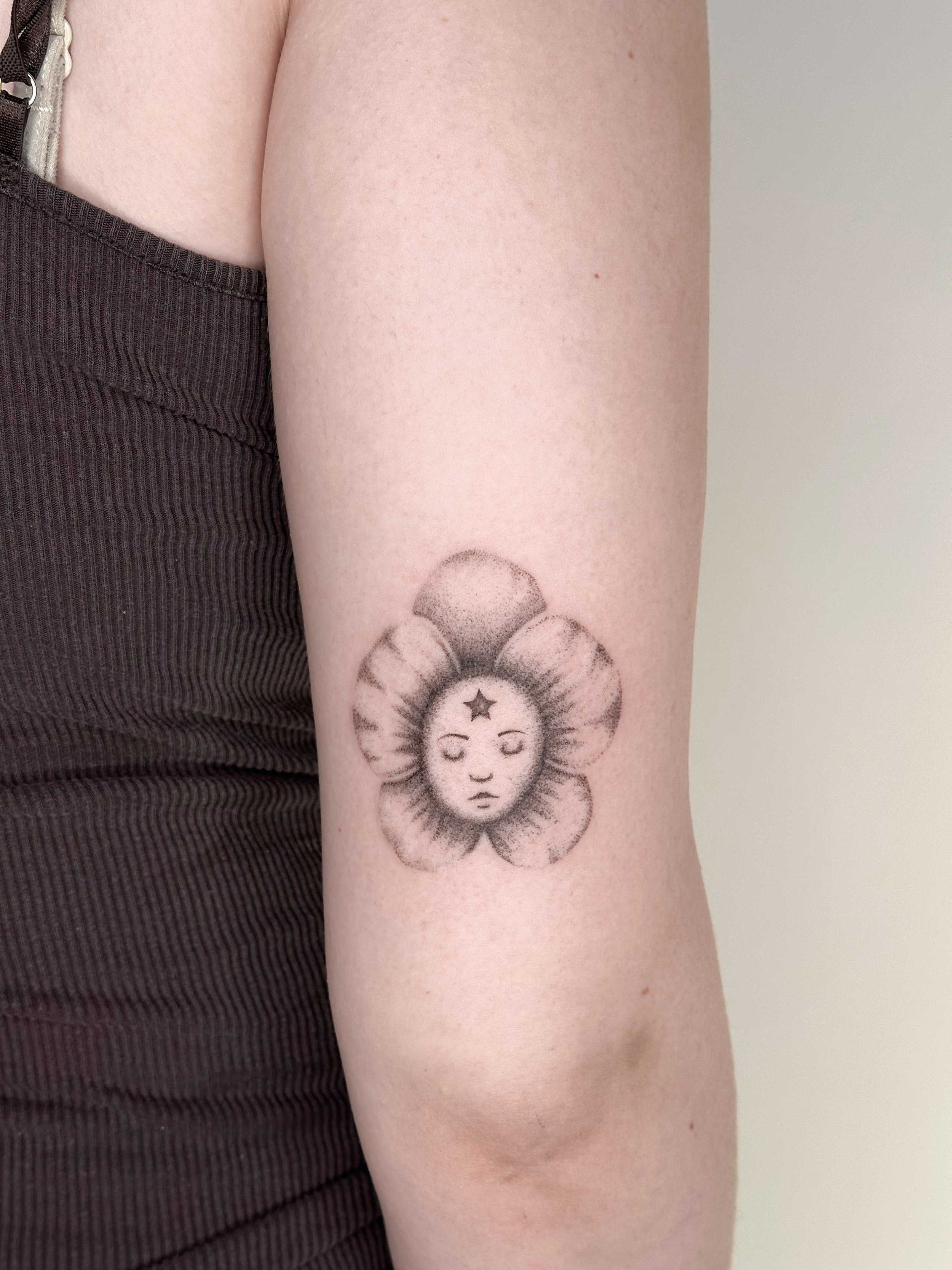 Black and gray tattoo of a woman's face with closed eyes, a star on the forehead, and flower petals surrounding her face, on person's upper arm.