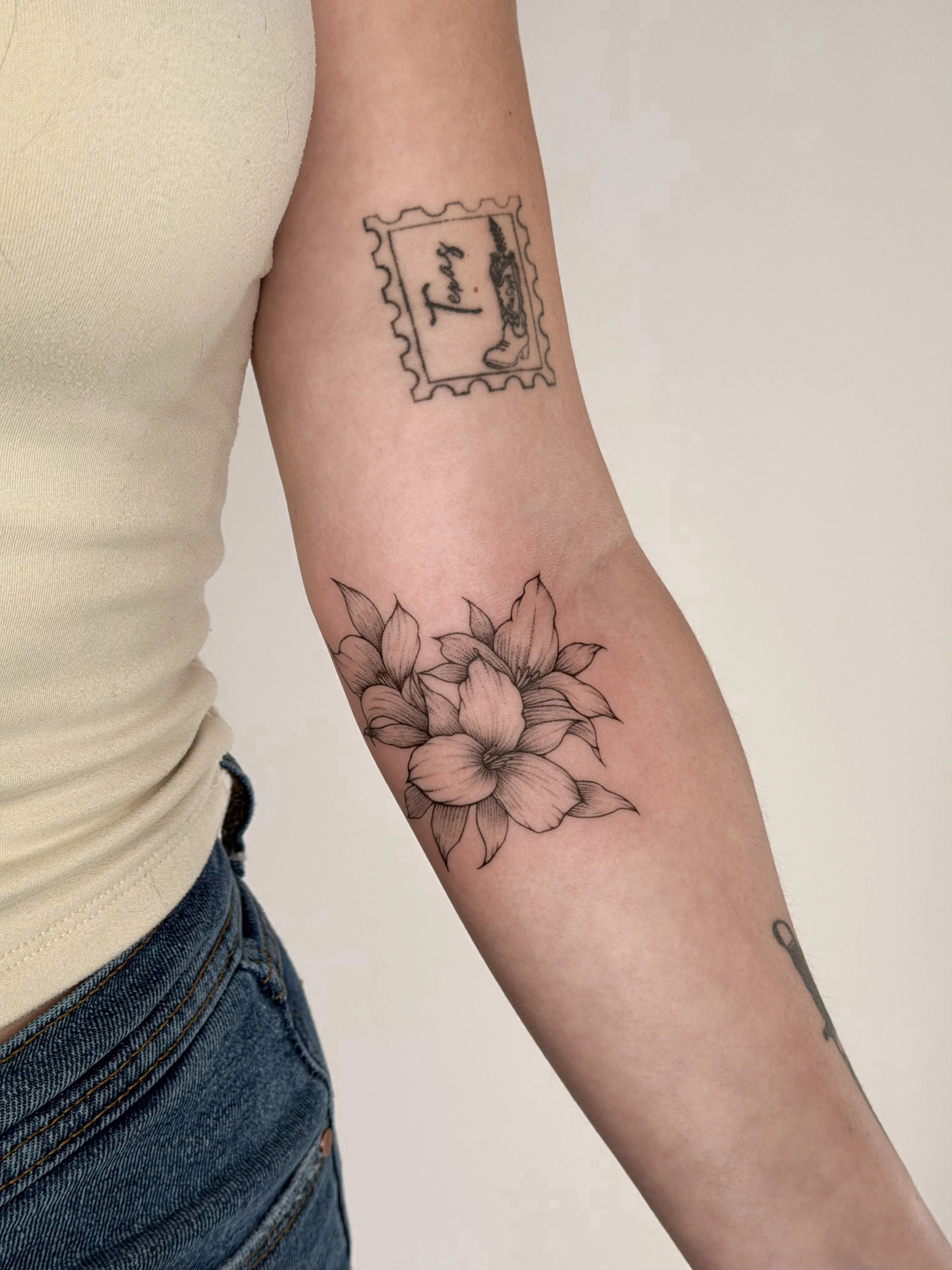 Tattoo of a flower with detailed petals on a person's forearm and a tattoo of a postage stamp with the word 'Tasty' and a signature on the upper arm.