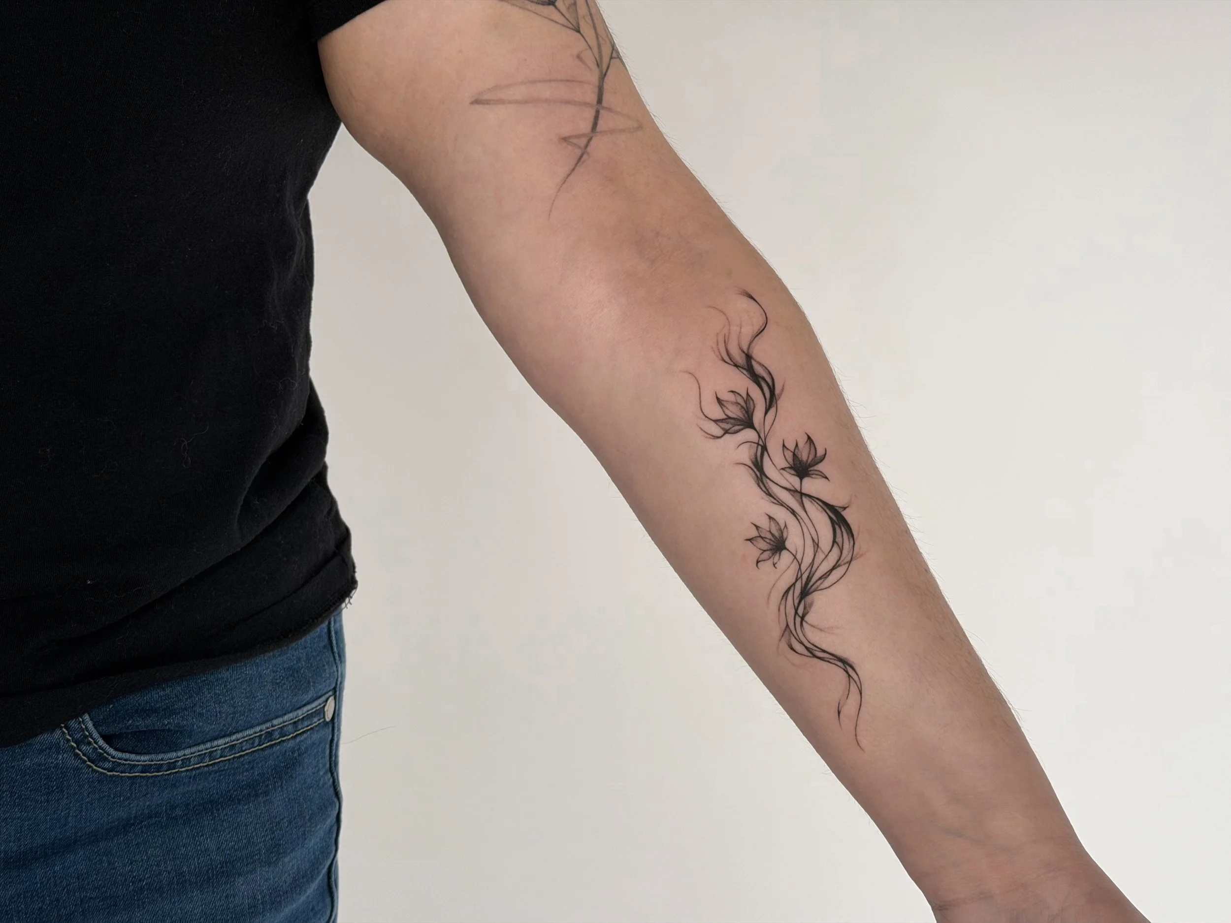 A person's arm with a black tattoo of an abstract floral design featuring flowing lines and flowers.
