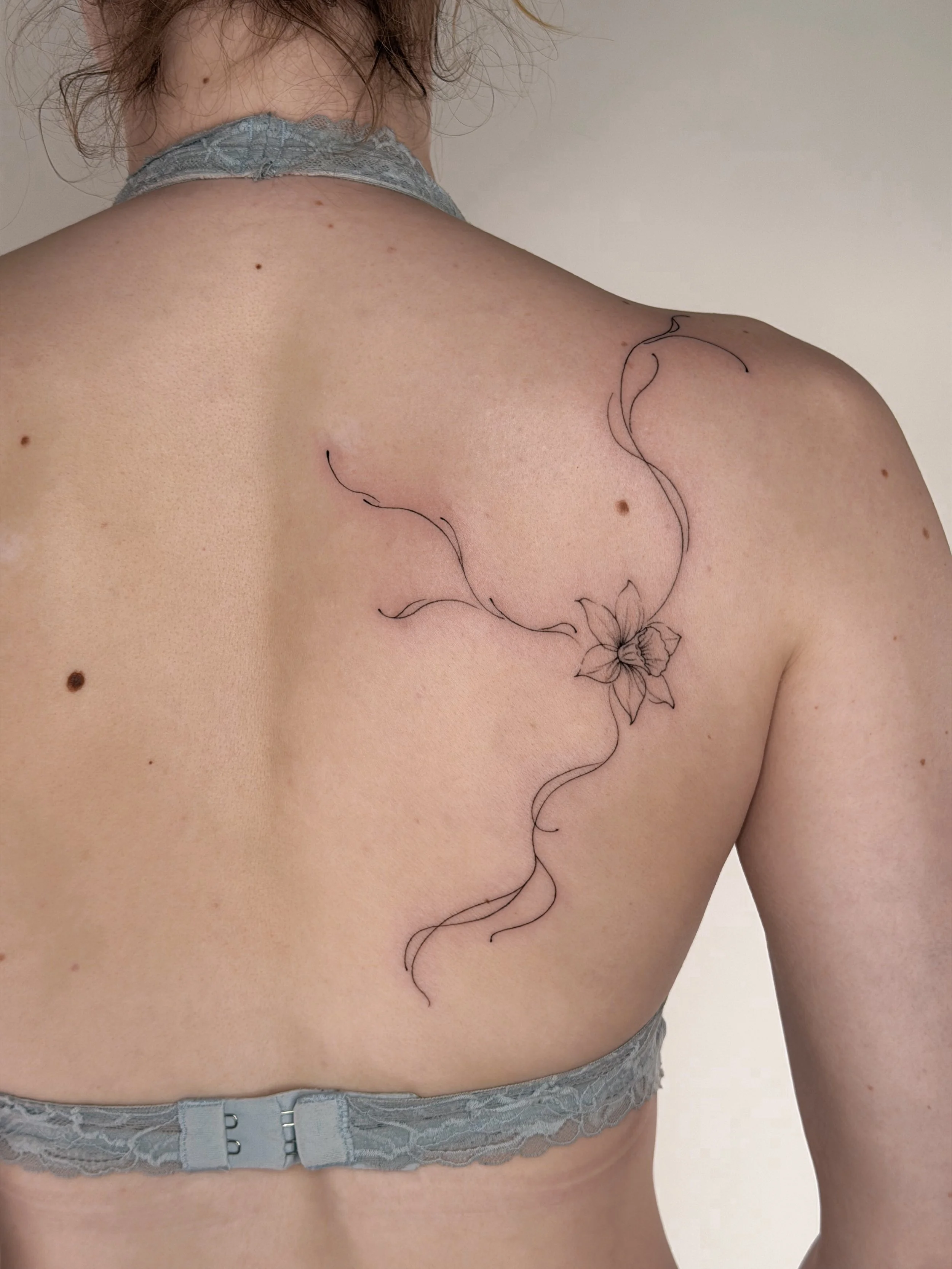 A close-up of a woman's back showing a tattoo of a flower with flowing lines extending from it.