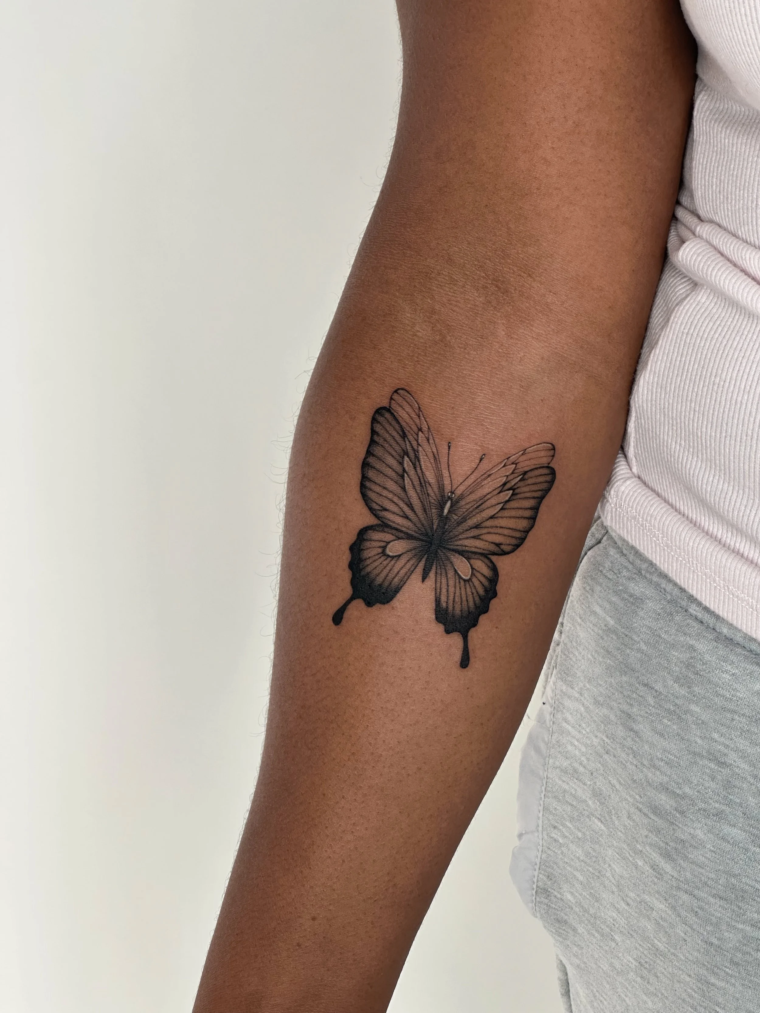 Black butterfly tattoo on person's forearm with white shirt and gray pants.