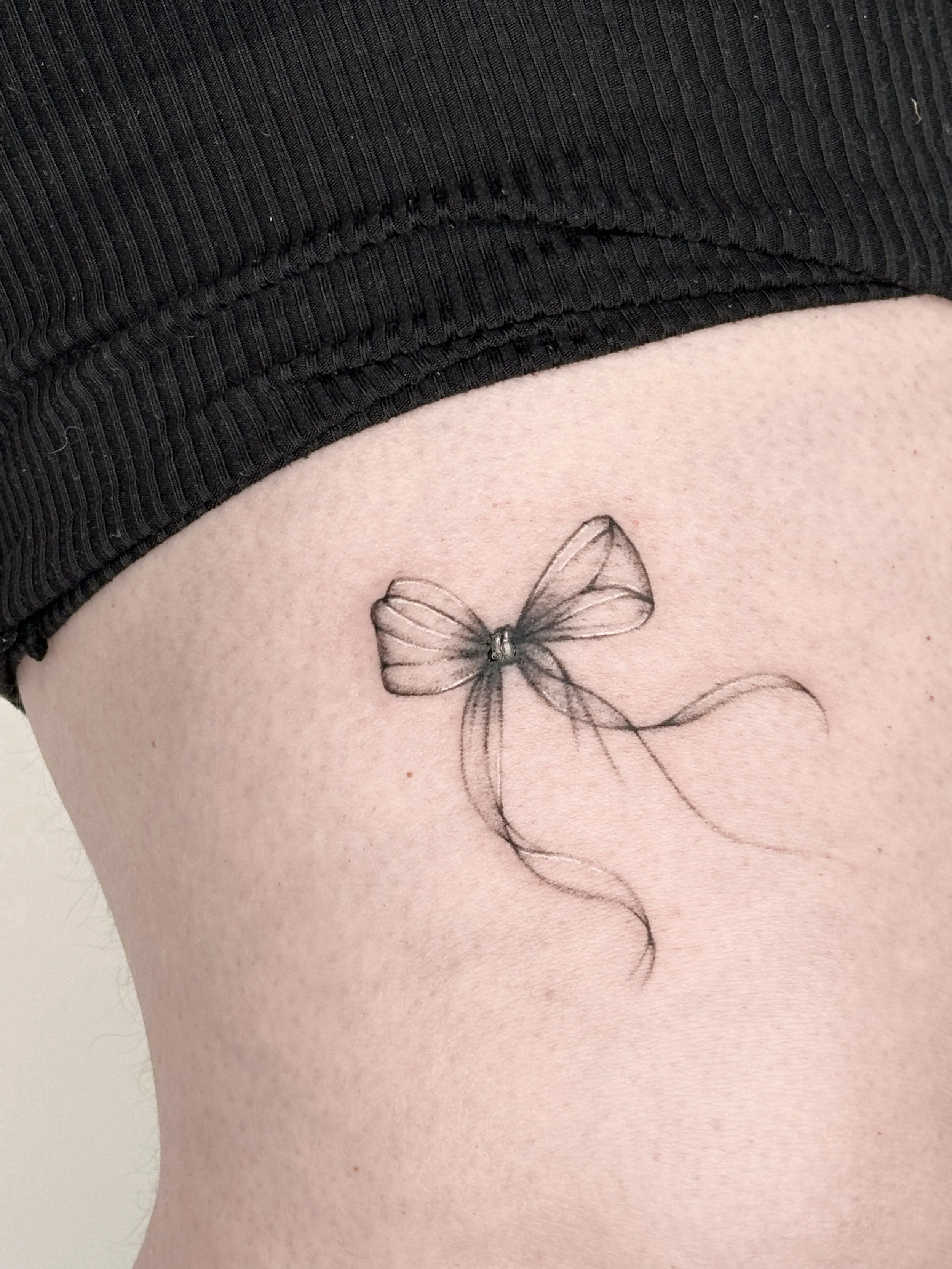 A delicate black ink tattoo of a bow with flowing ribbons on person's side near their waist.