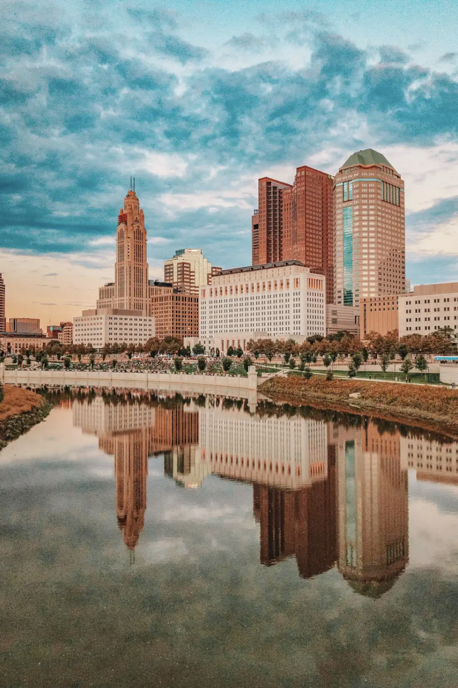 Skyline of Columbus, Ohio with skyscrapers reflecting in the Scioto River under a cloudy sky.