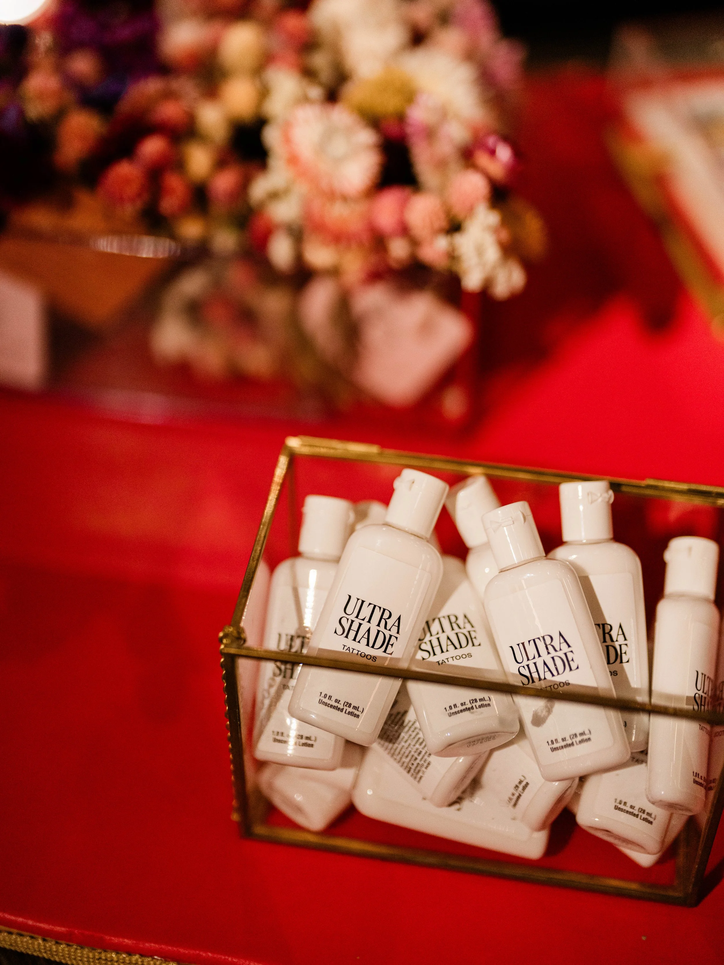 A collection of Ultra Shade tattoo lotion bottles in a glass container on a red table, with blurred flowers in the background.