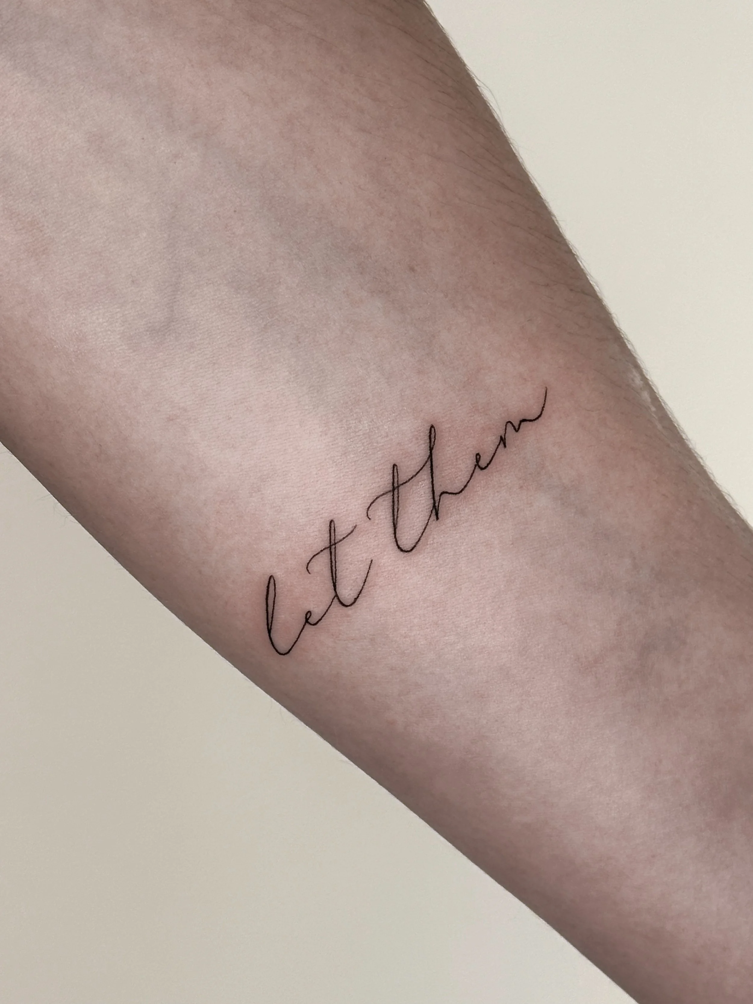 Tattoo on a person's arm that says 'let them' in cursive script.