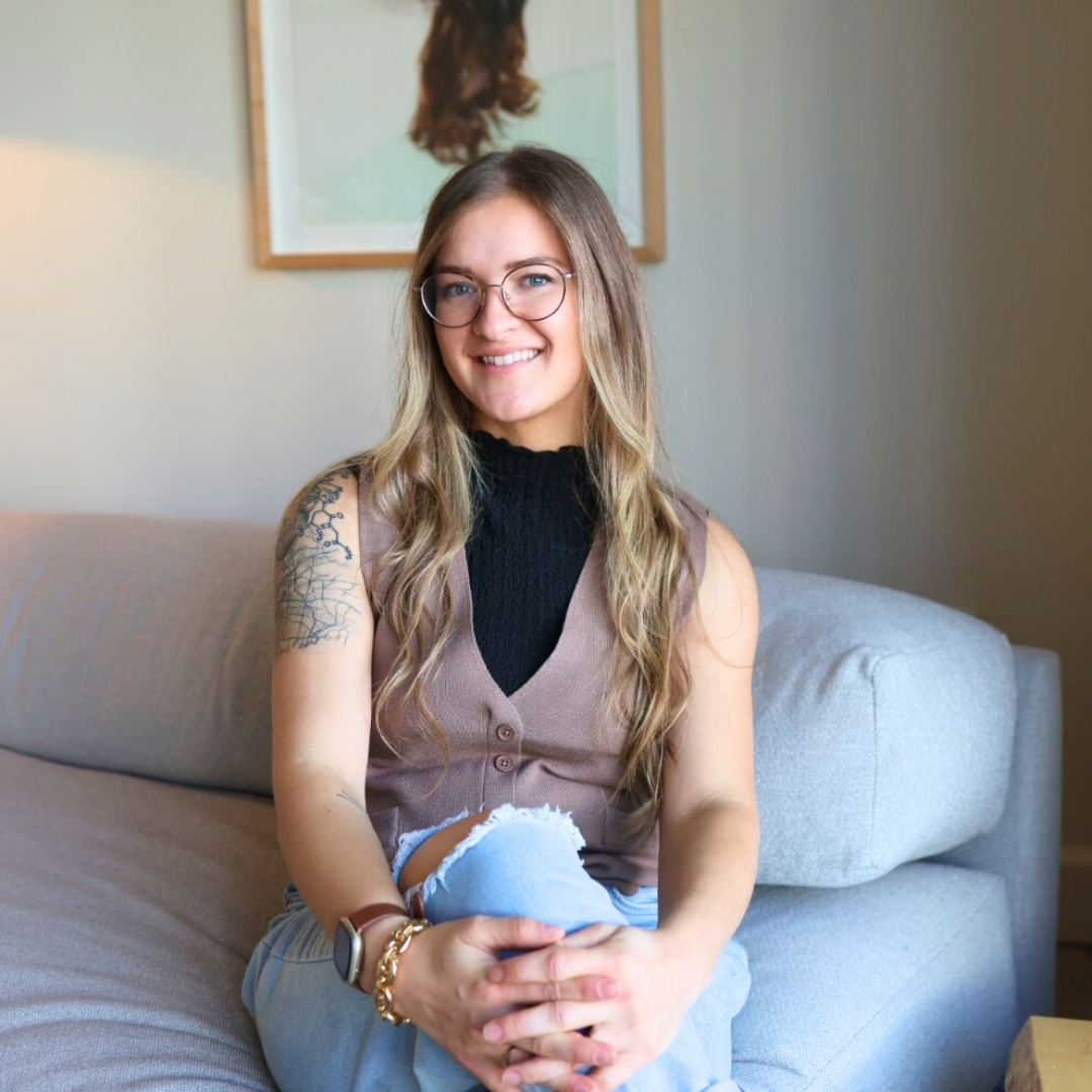 Woman with glasses sitting on a couch indoors, wearing a vest and ripped jeans, with a tattoo on her arm.