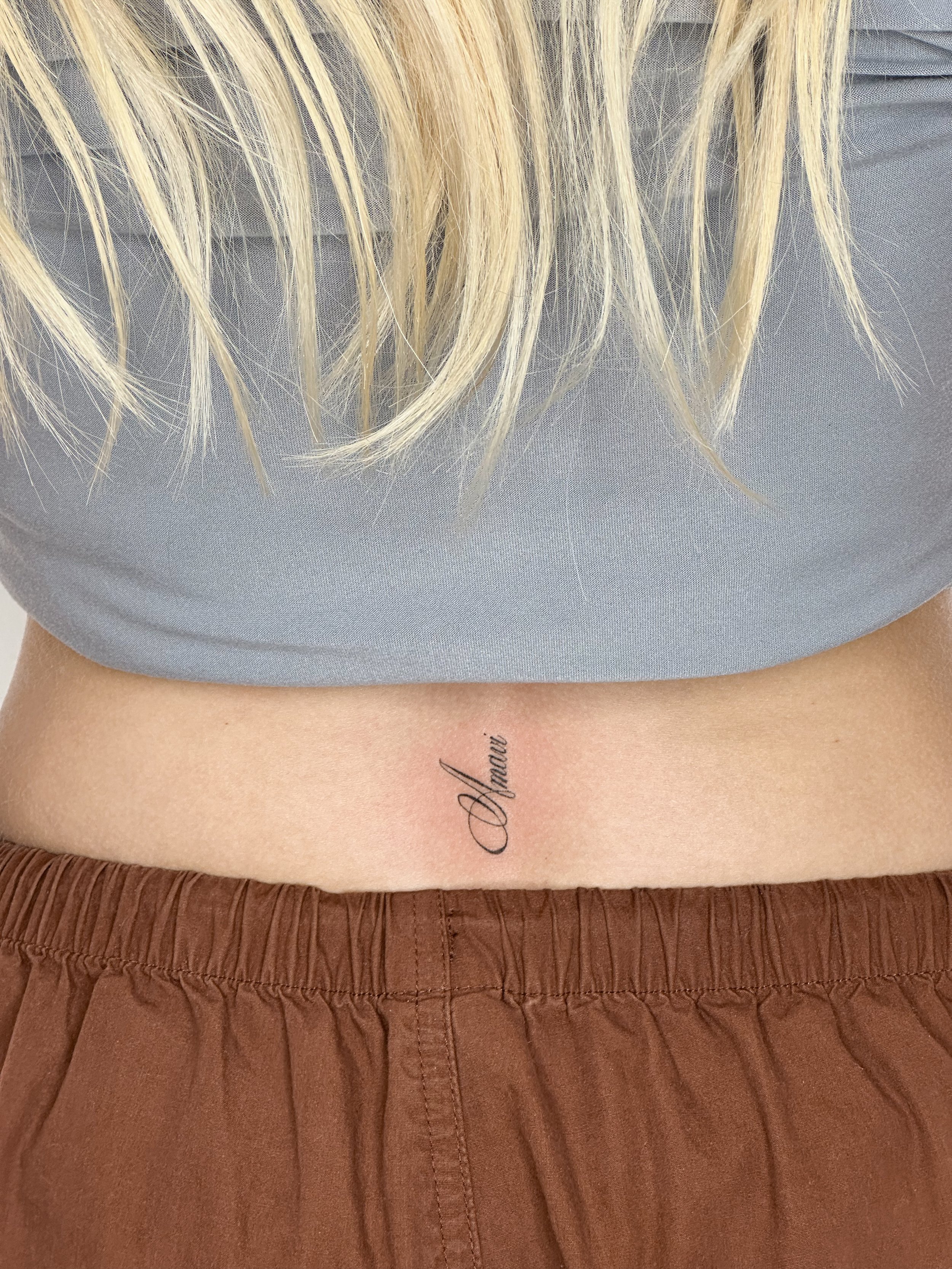 Tattoo on lower back with the word 'Amavi' in cursive script, below blonde hair, wearing a gray top and brown pants.