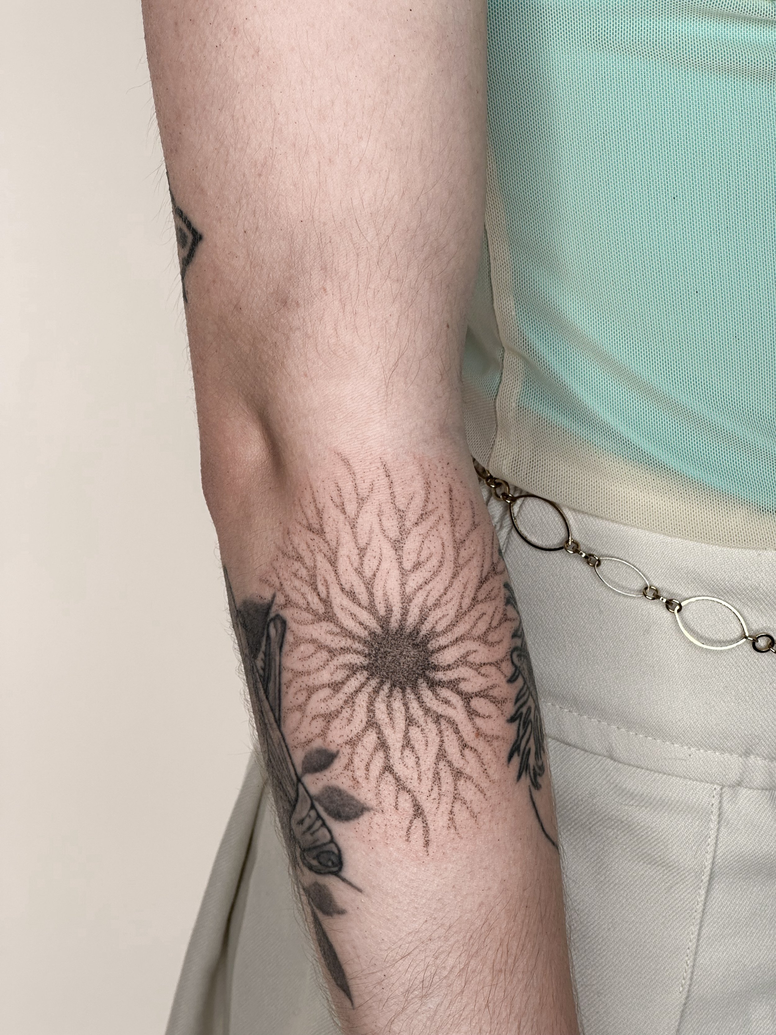 Close-up of a person's arm with a sunburst design tattoo and a partial view of other tattoos. A person is wearing light-colored clothing with metal chain accents.